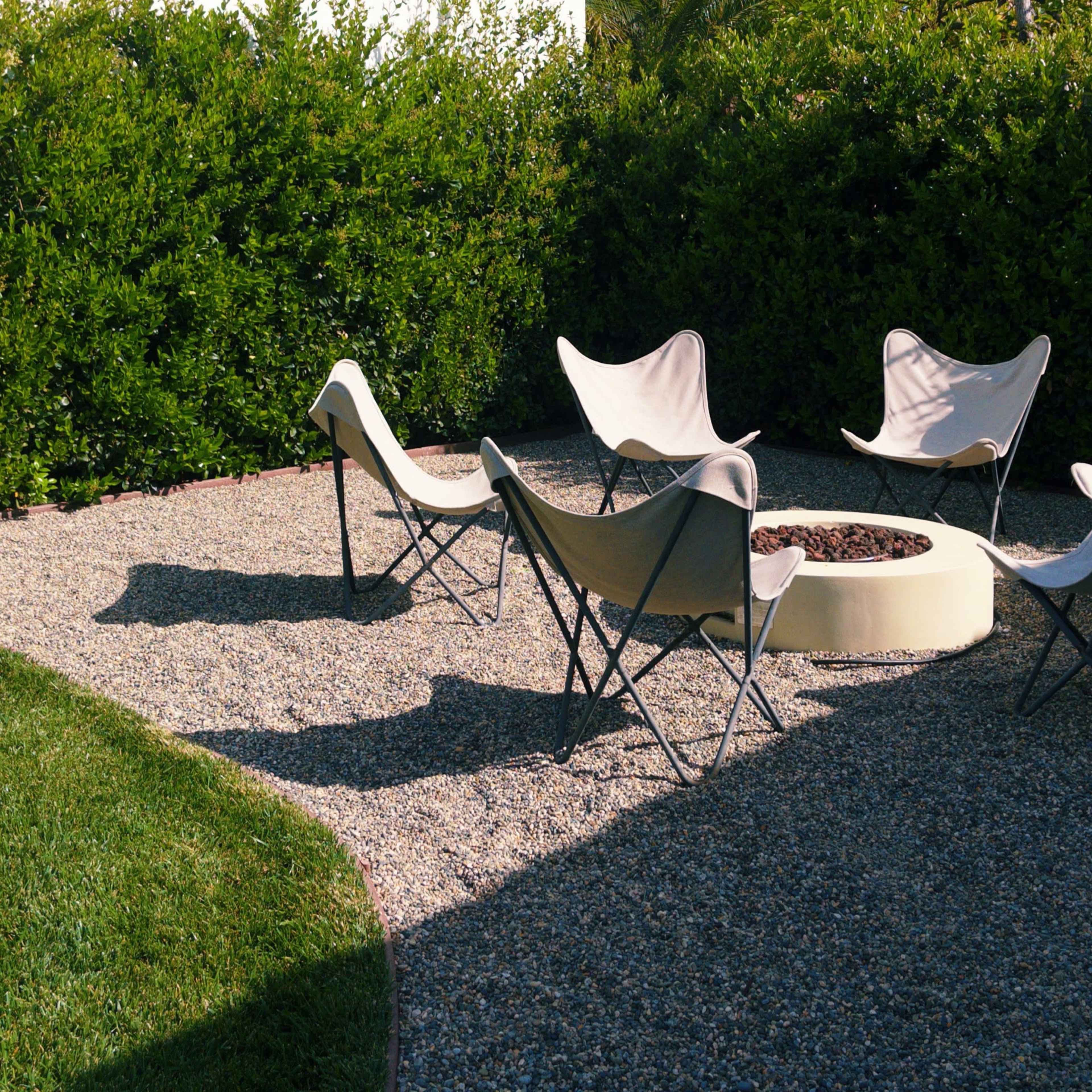 A gravel patio featuring five lounge chairs arranged around a fire pit, surrounded by green shrubs.
