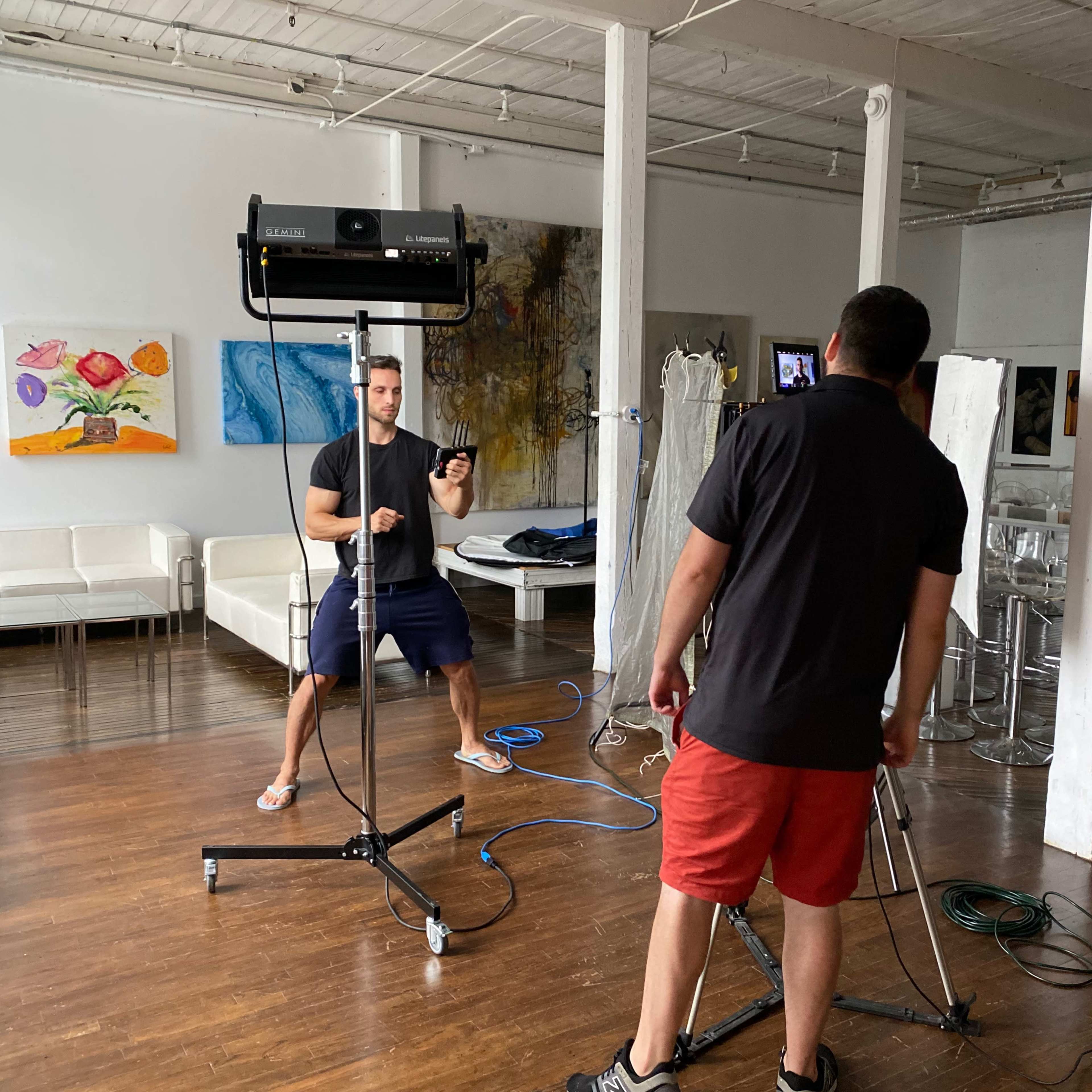 Two men are setting up lighting equipment in a spacious studio with artwork on the walls.