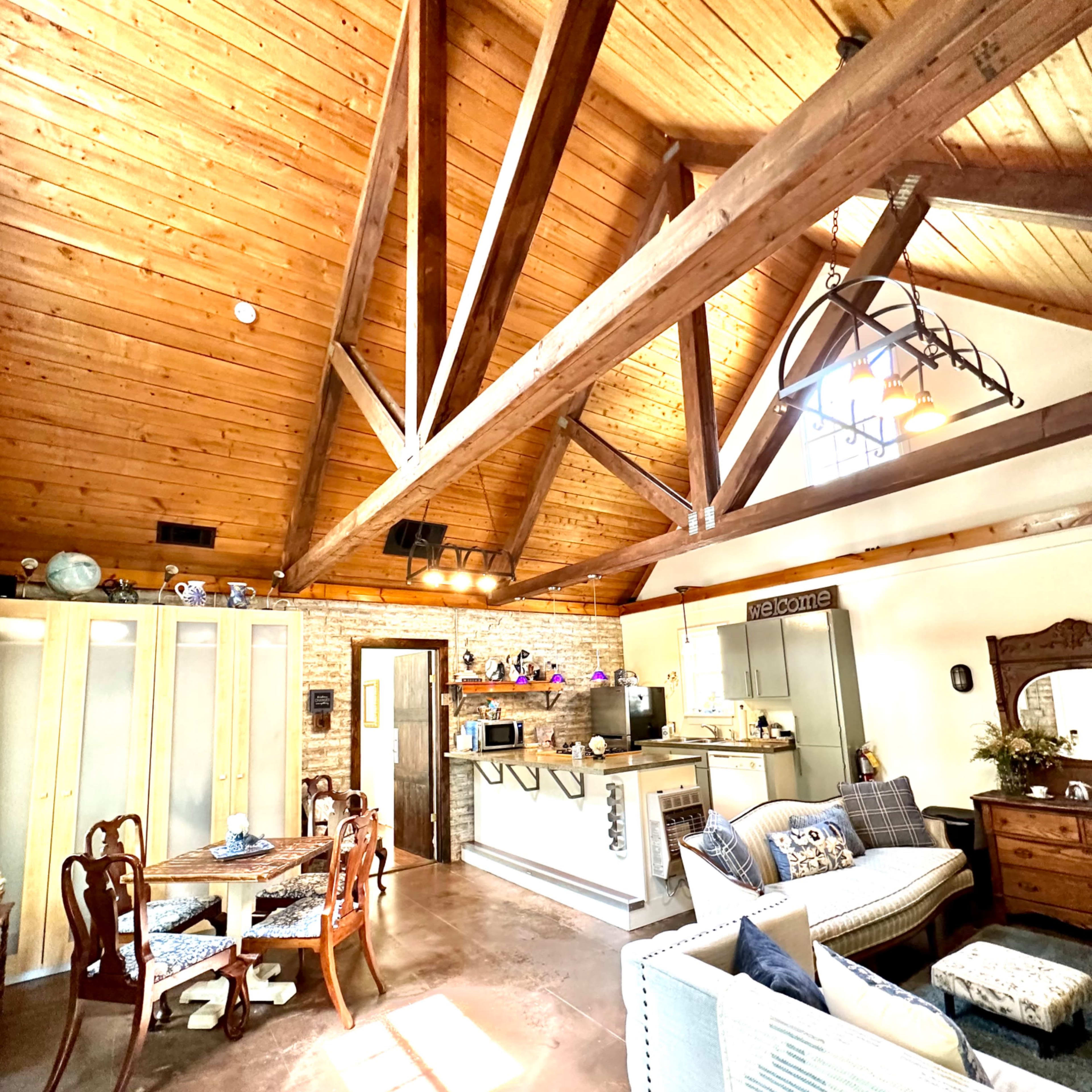 A spacious, rustic-style living area with a high wooden beam ceiling, a dining table, a sofa, and a kitchen area in the background.