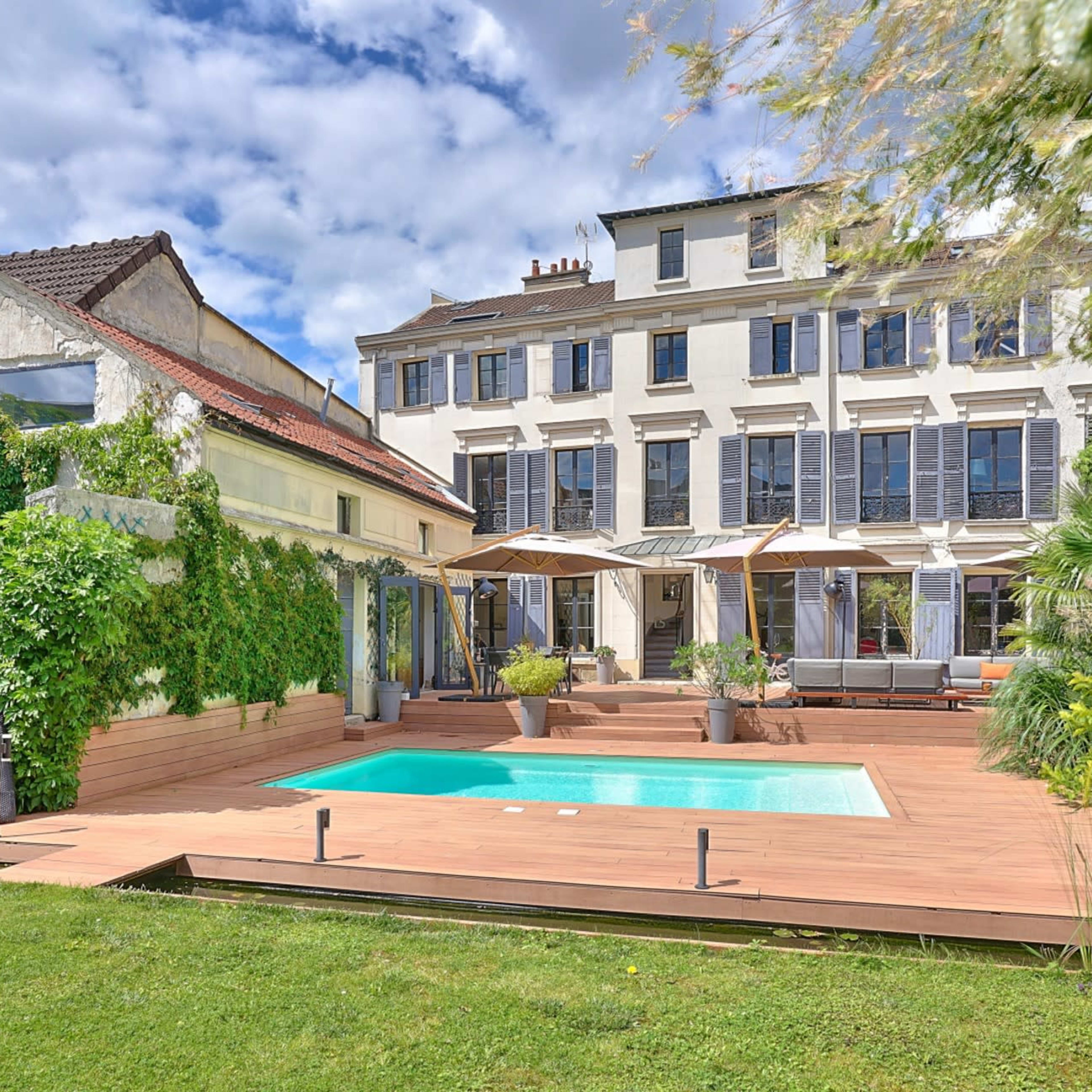 A landscaped backyard featuring a swimming pool, surrounded by a wooden deck and a two-story house with multiple windows and patio umbrellas.