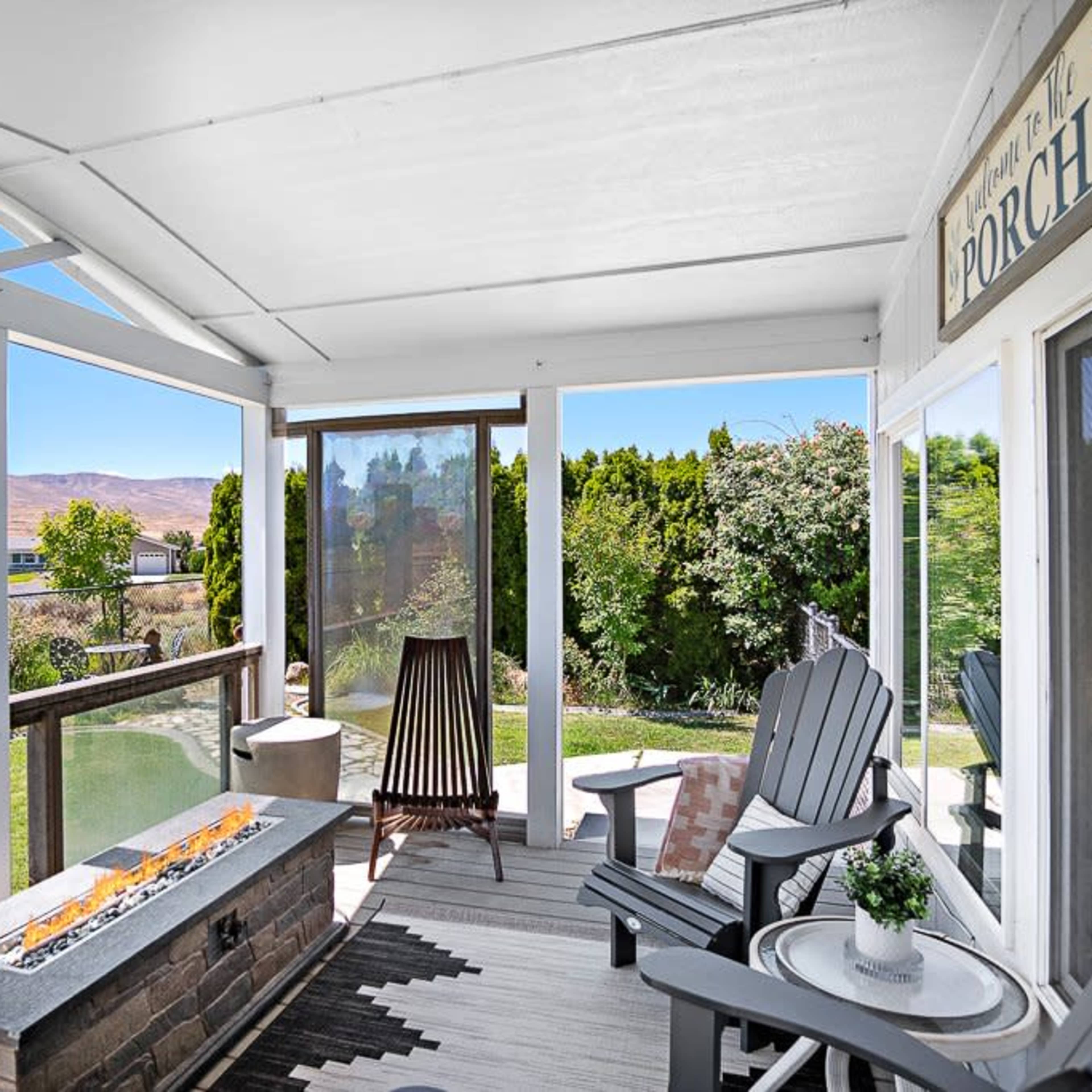 A covered porch features a stone fire pit, adirondack chairs, and views of a landscaped yard and distant mountains.