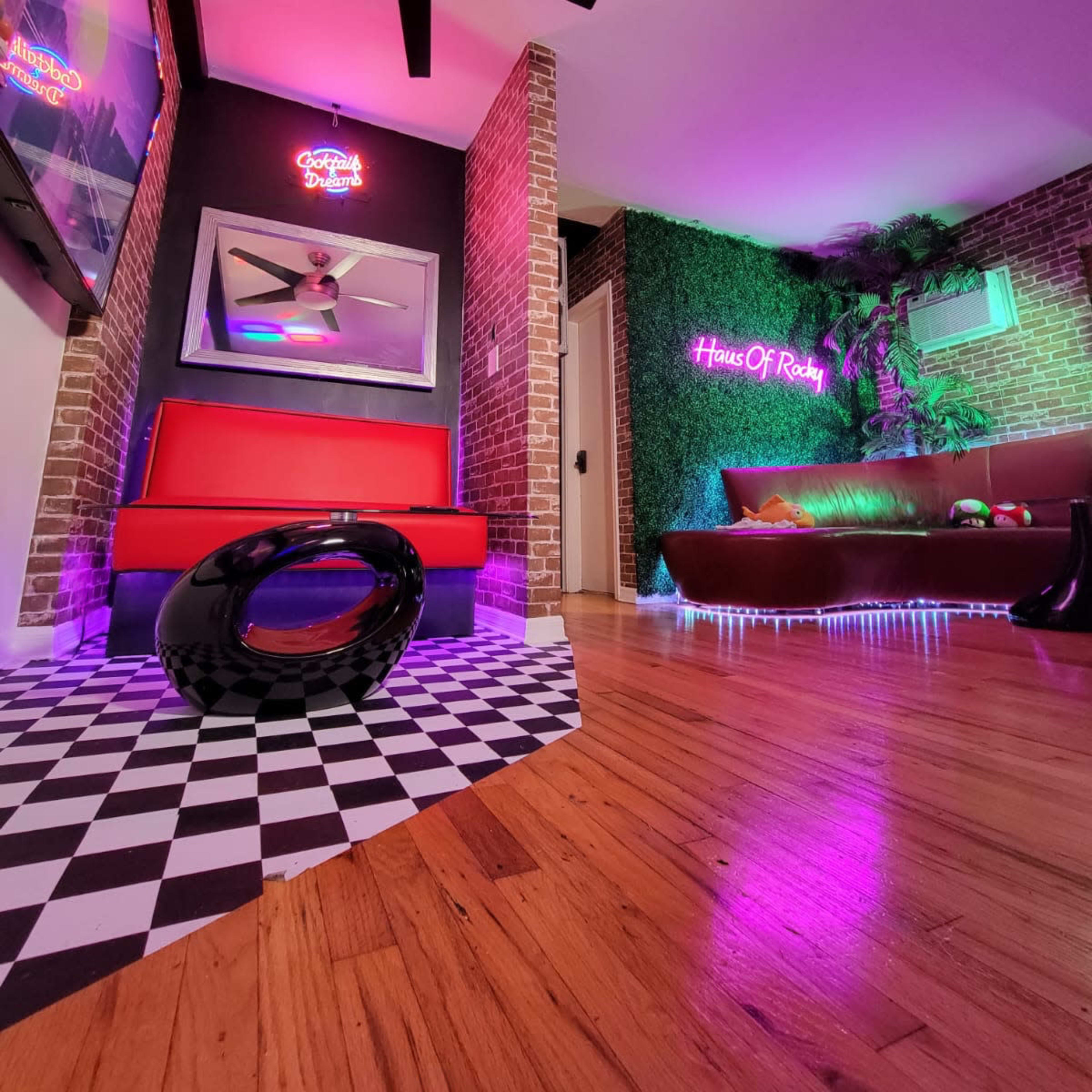 A modern room featuring a red couch, a dark brown sofa, and colorful neon lights against a brick wall and green foliage.