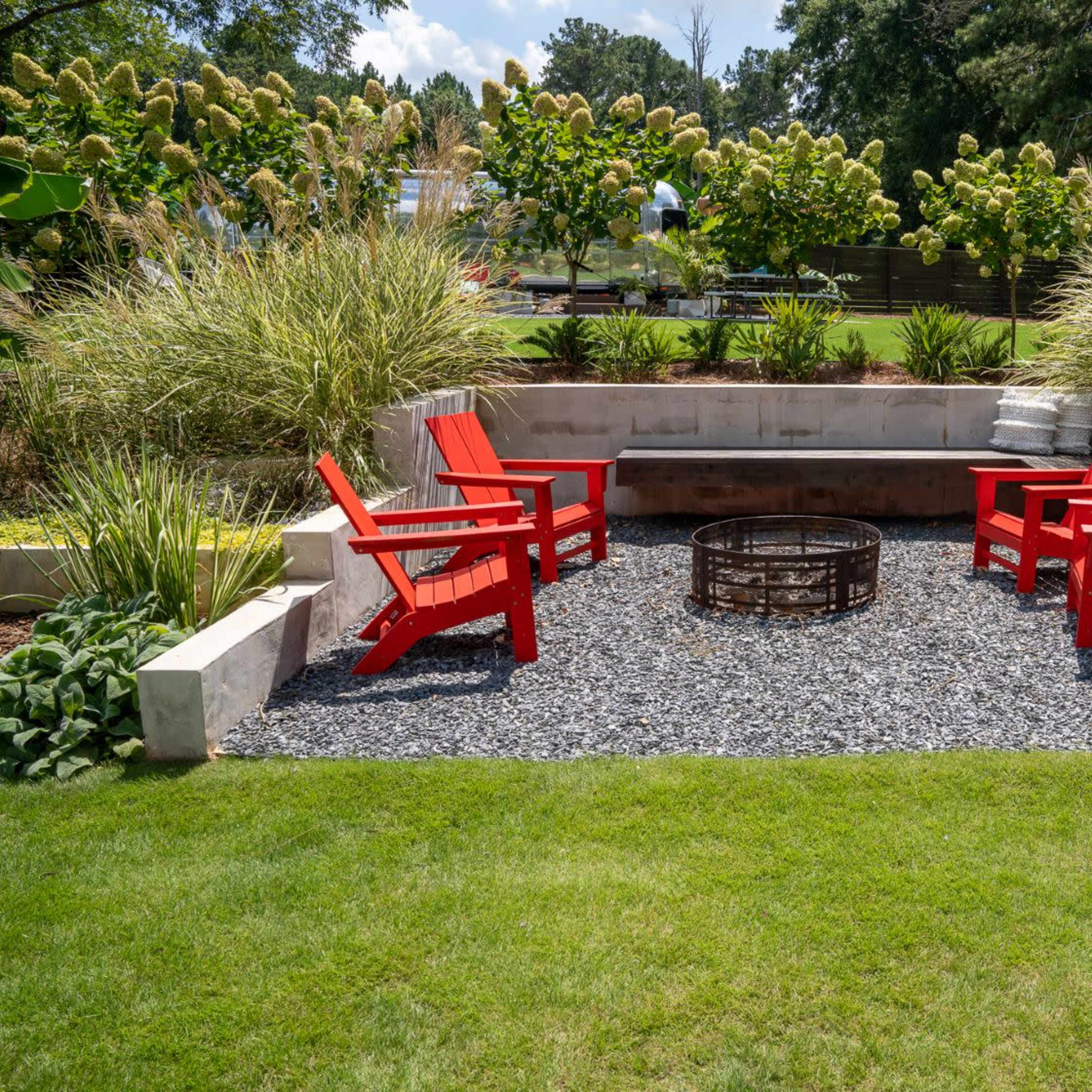 A fire pit is surrounded by four red Adirondack chairs on a gravel area within a landscaped garden.