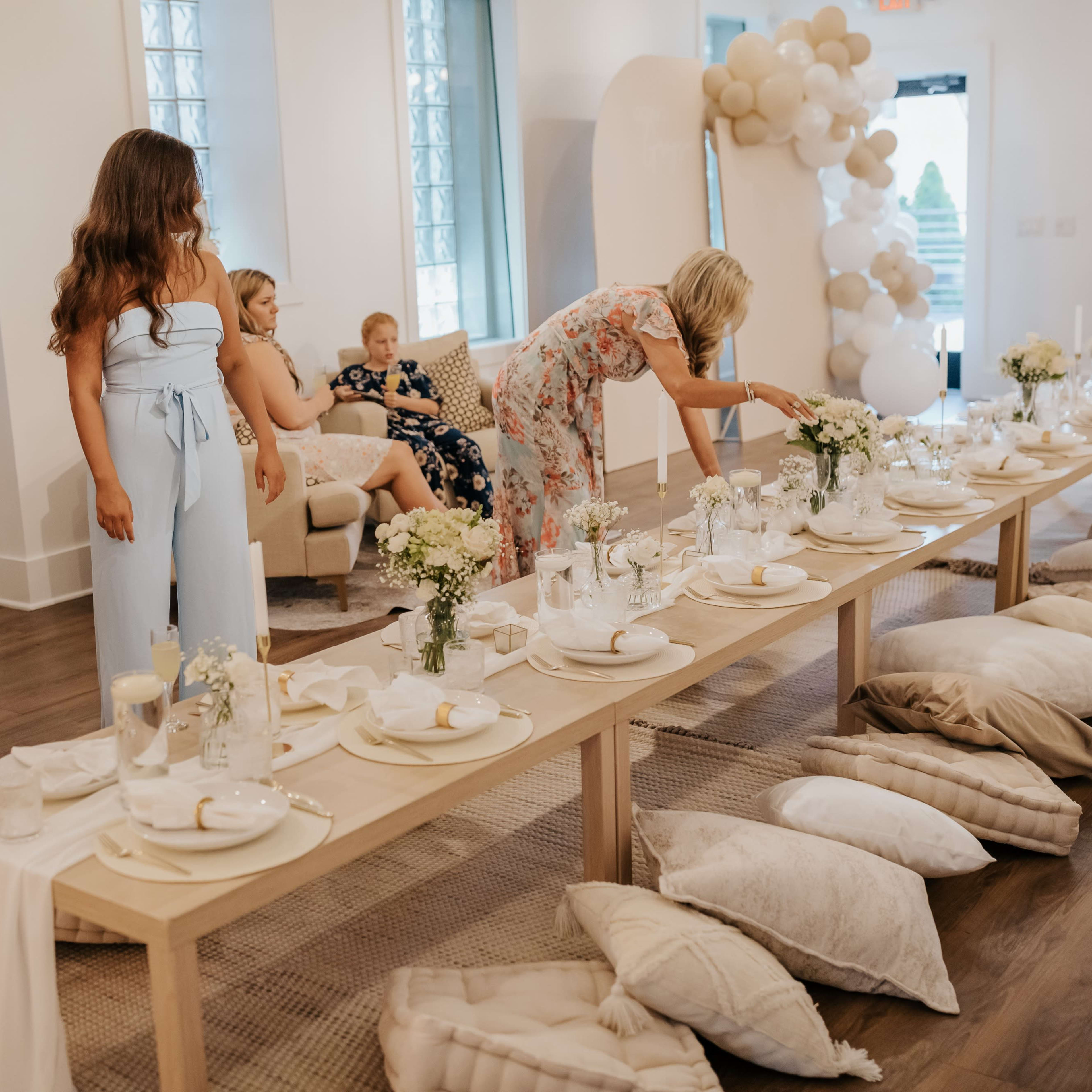A long table is elegantly set with plates and flowers, surrounded by guests in a decorated venue featuring light-colored walls and a balloon arch.