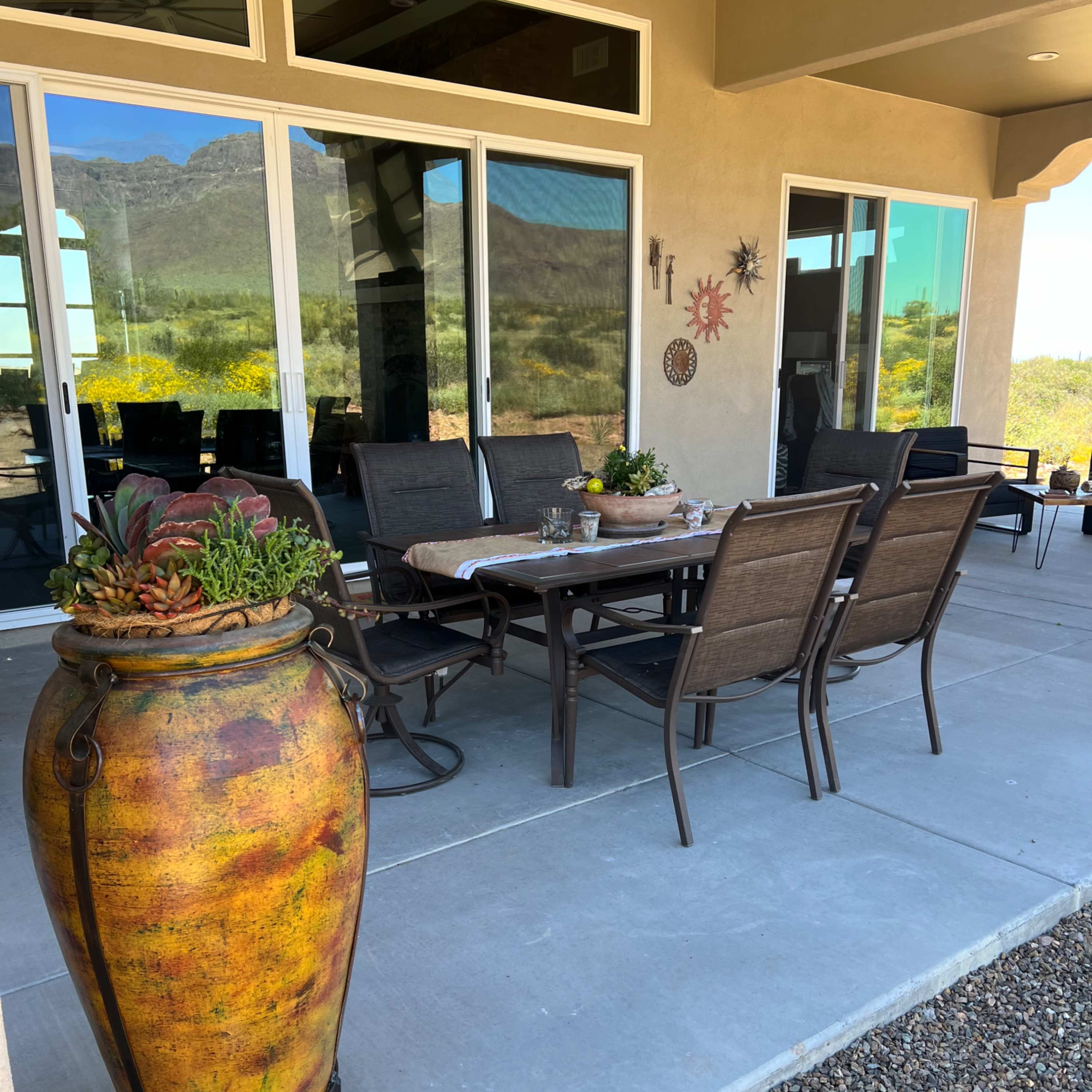A patio area features a dining table surrounded by chairs, complemented by a large decorative pot and sliding glass doors that overlook a mountainous landscape.