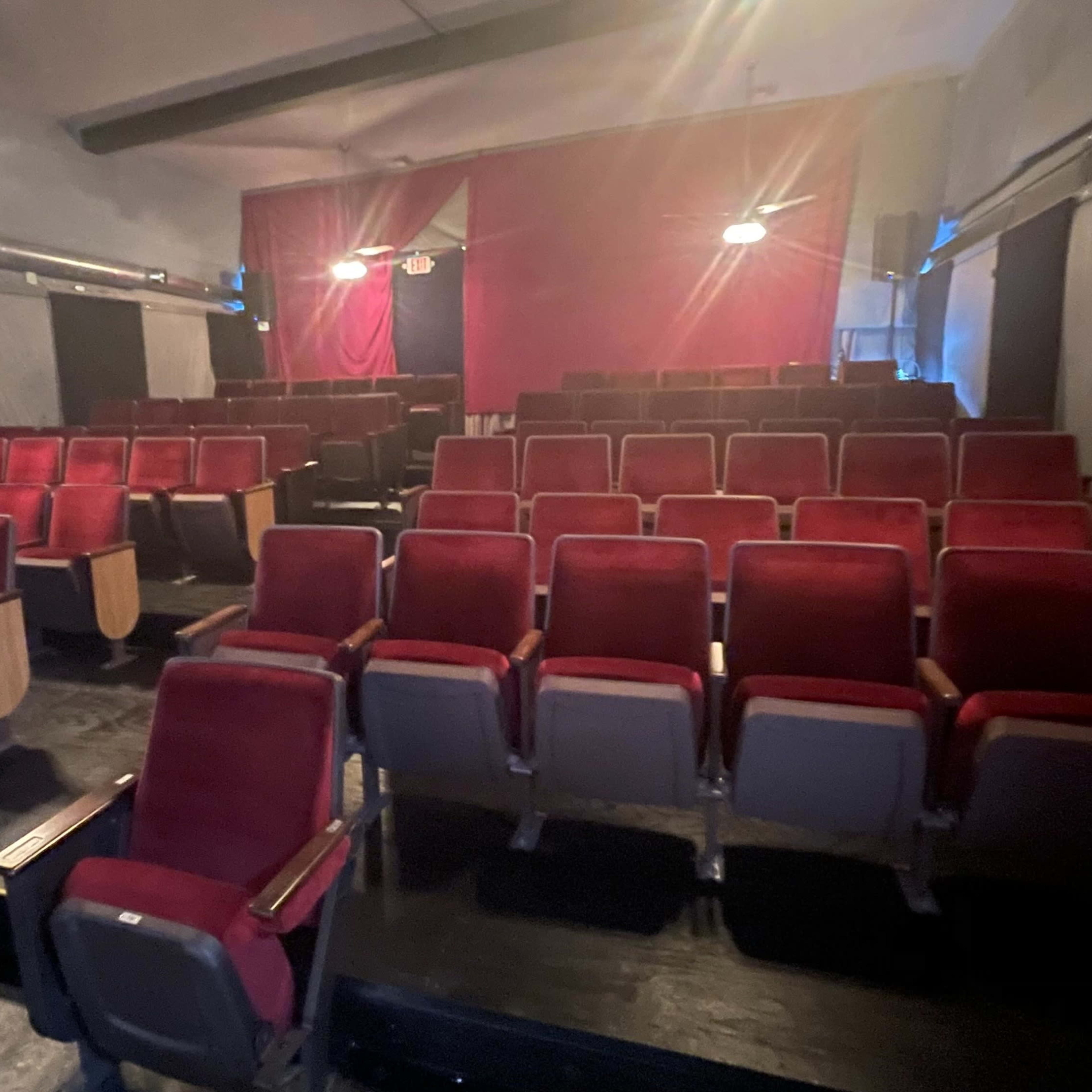 An empty theater seating area with rows of red chairs arranged in front of a stage draped in fabric.