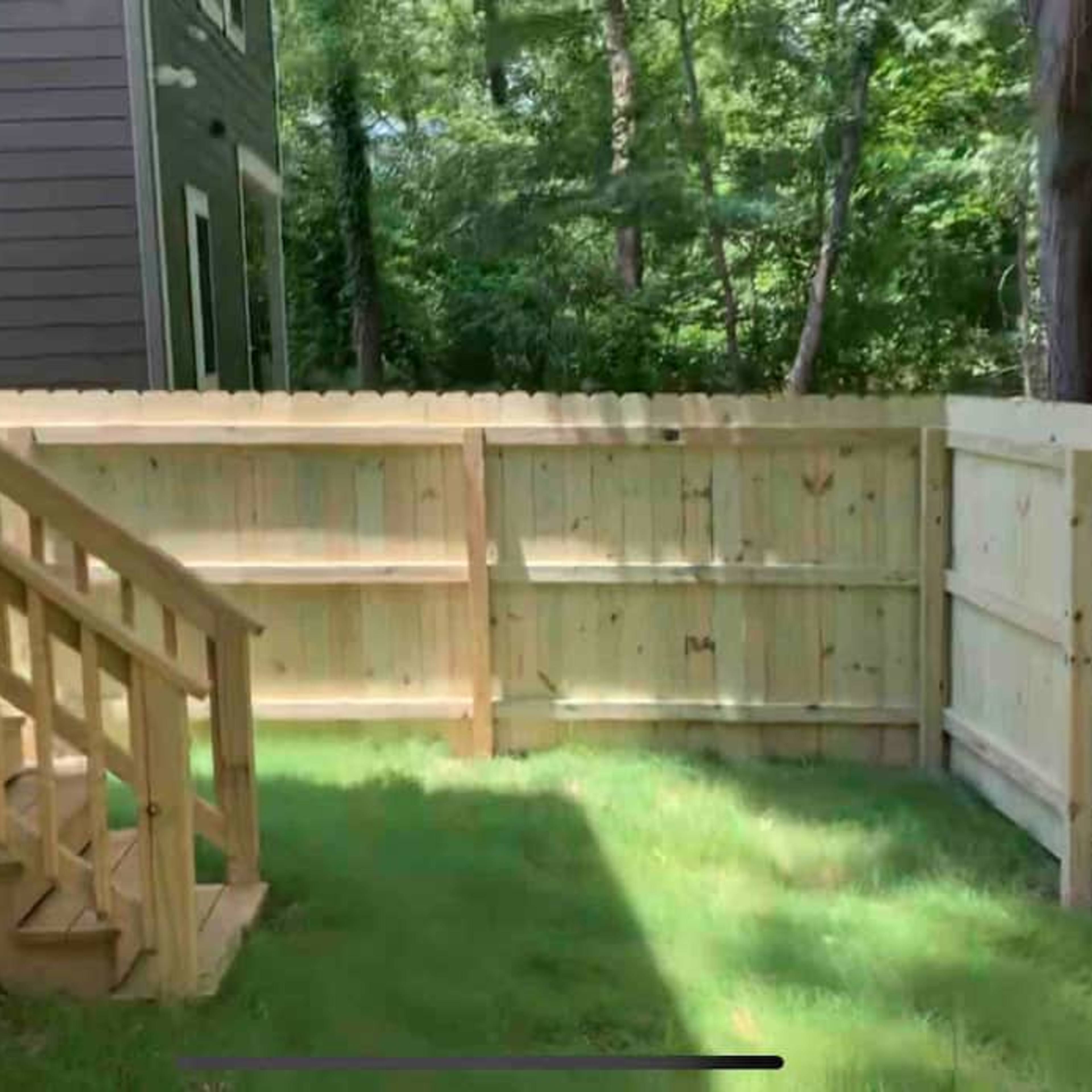 A wooden staircase leads from a house to a fenced-in backyard with green grass and trees in the background.