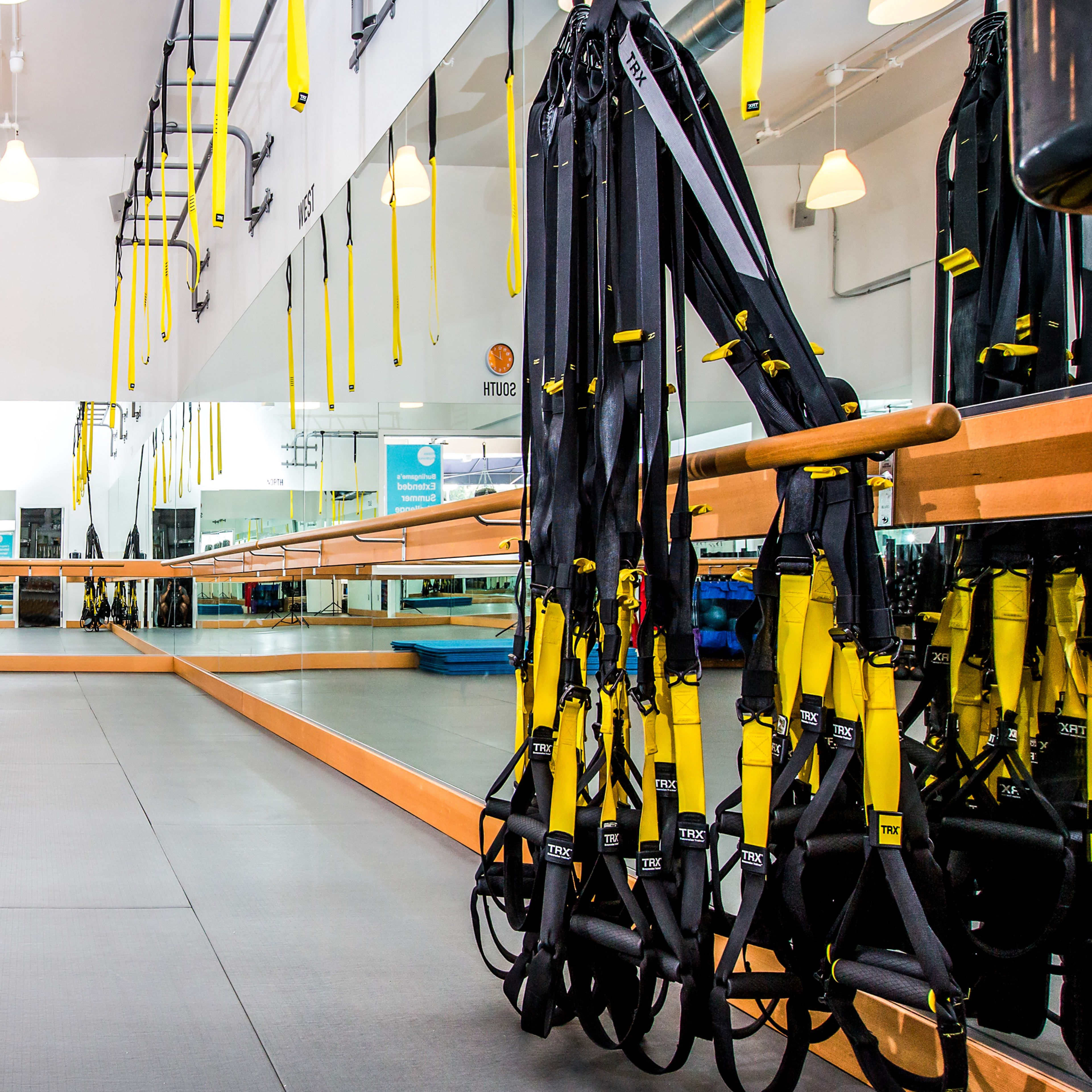 A gym interior equipped with suspension training straps hanging from a mirrored wall, with workout mats and exercise equipment in the background.