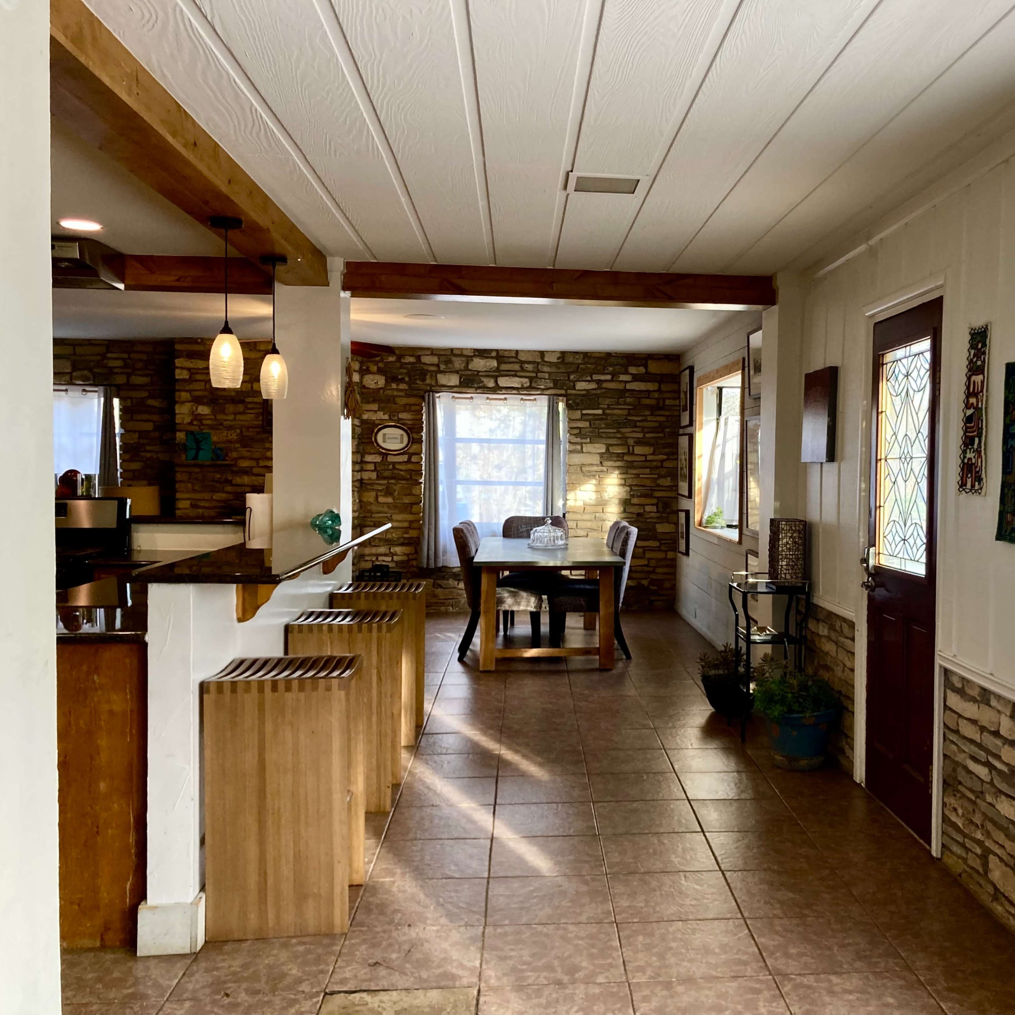 A spacious interior with a dining area featuring a round table and chairs, and a kitchen bar with wooden stools, all set against a backdrop of stone walls and natural light from windows.