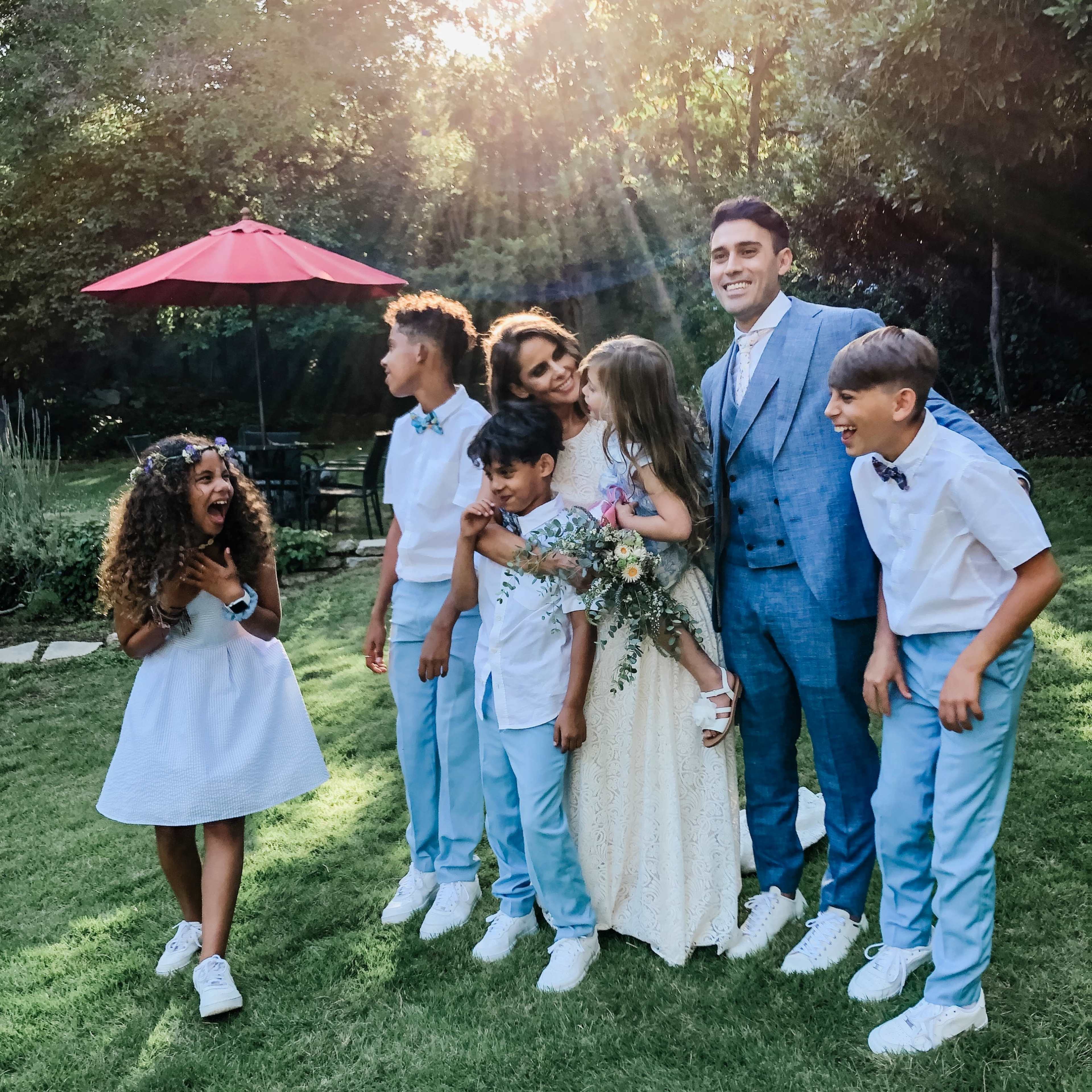 A family poses together in a sunny garden, with a bride and groom standing at the center surrounded by children.