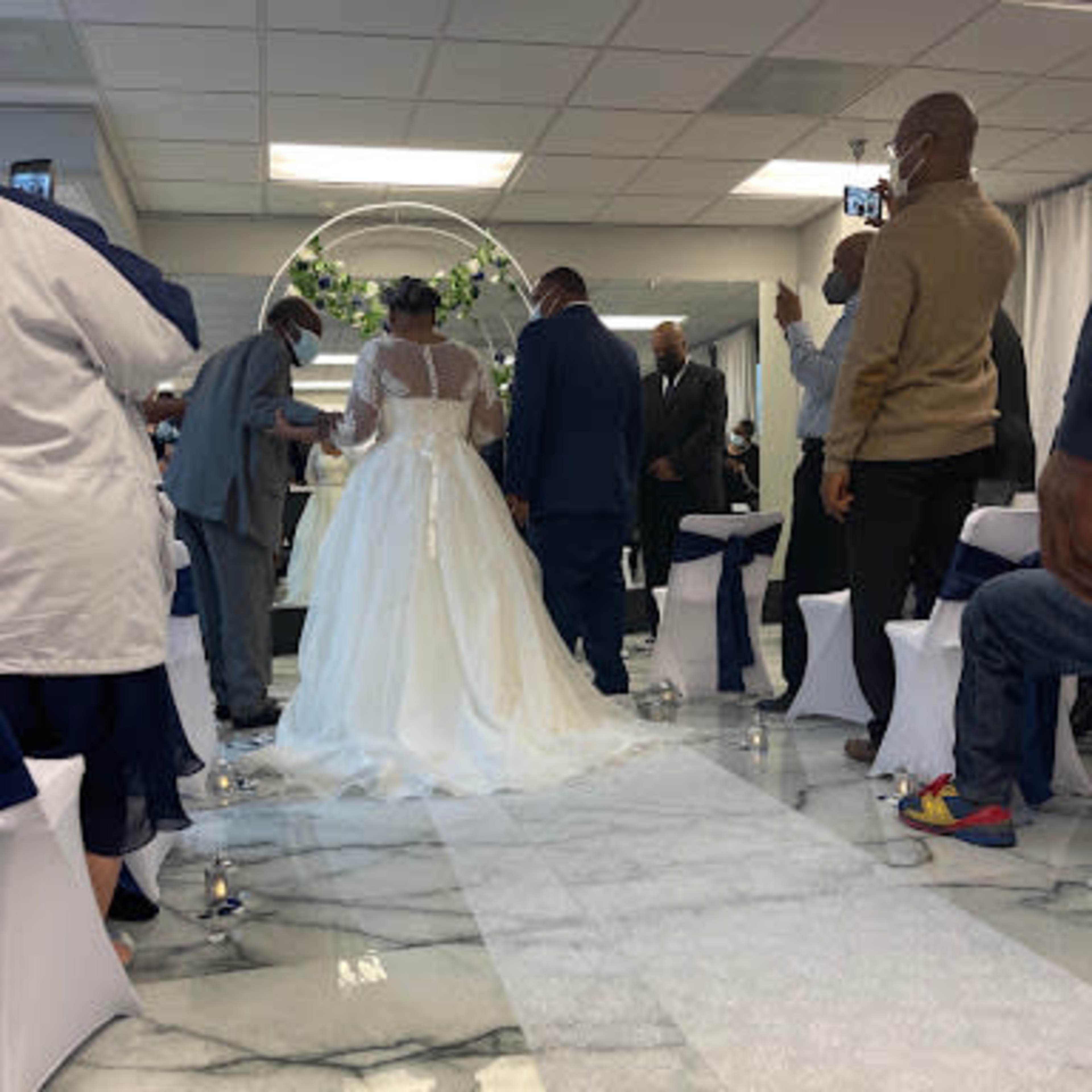 A bride in a white gown walks down a floral-decorated aisle surrounded by guests in formal attire.