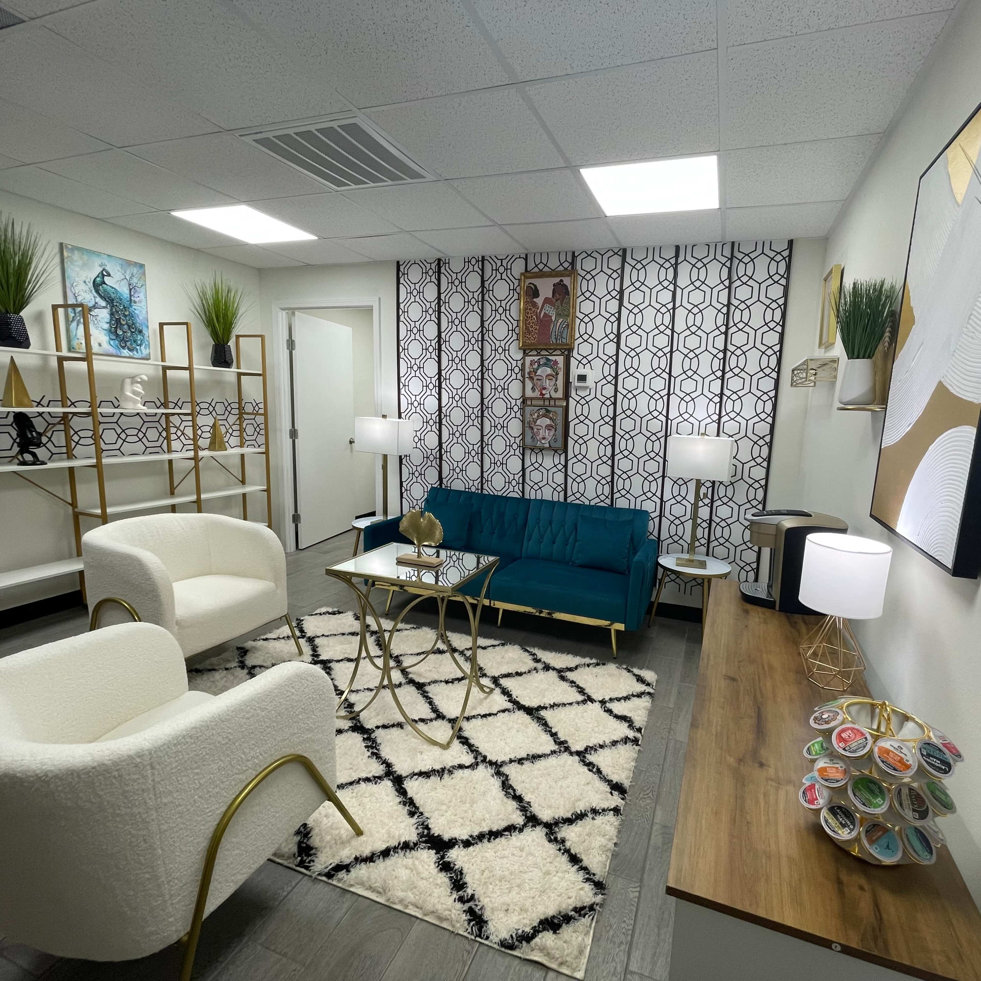 A modern lounge area with a teal sofa, two white armchairs, a coffee table, and decorative plants on shelves against a patterned wall.
