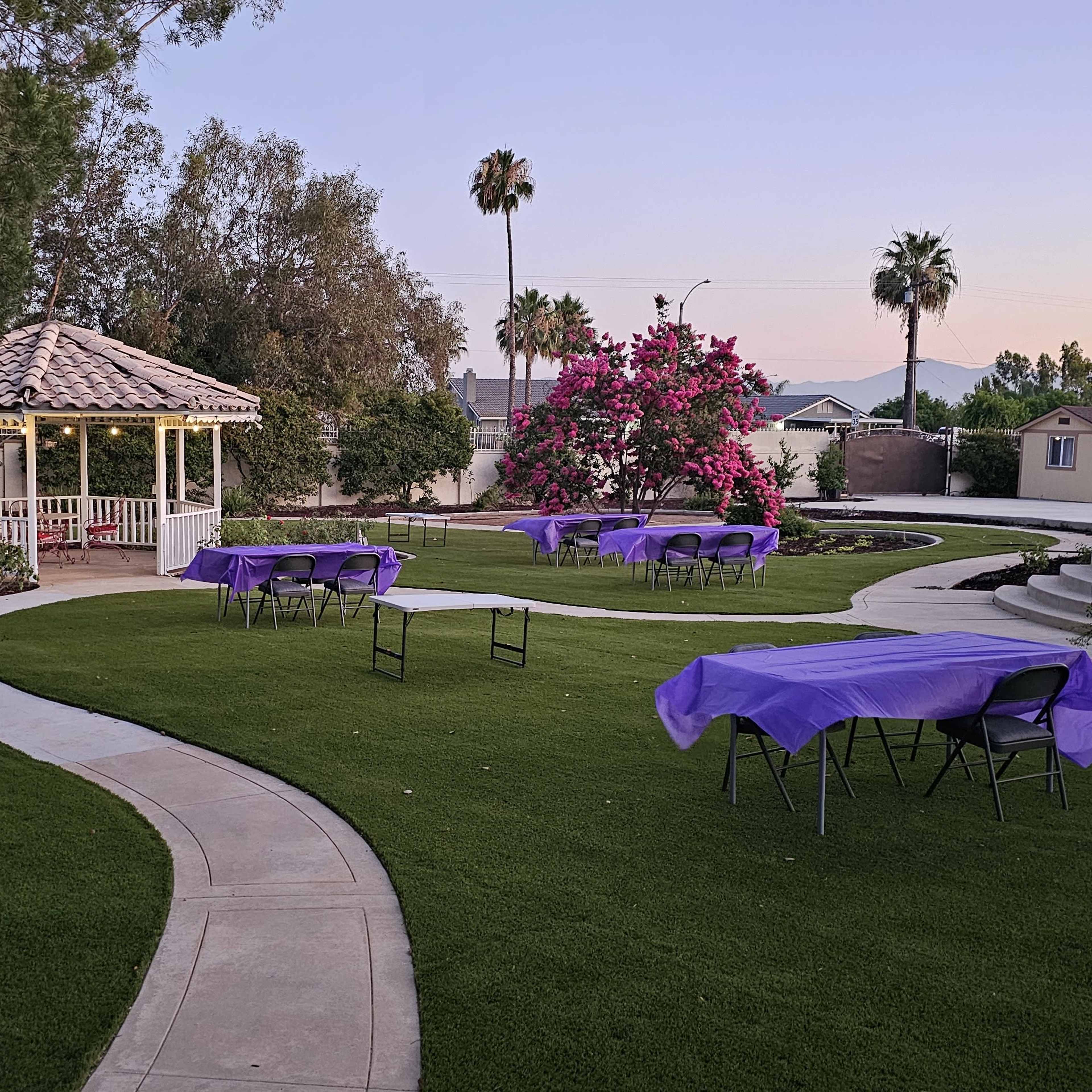 A landscaped area with a gazebo, winding pathways, and several tables covered with purple tablecloths surrounded by greenery and blooming flowers.
