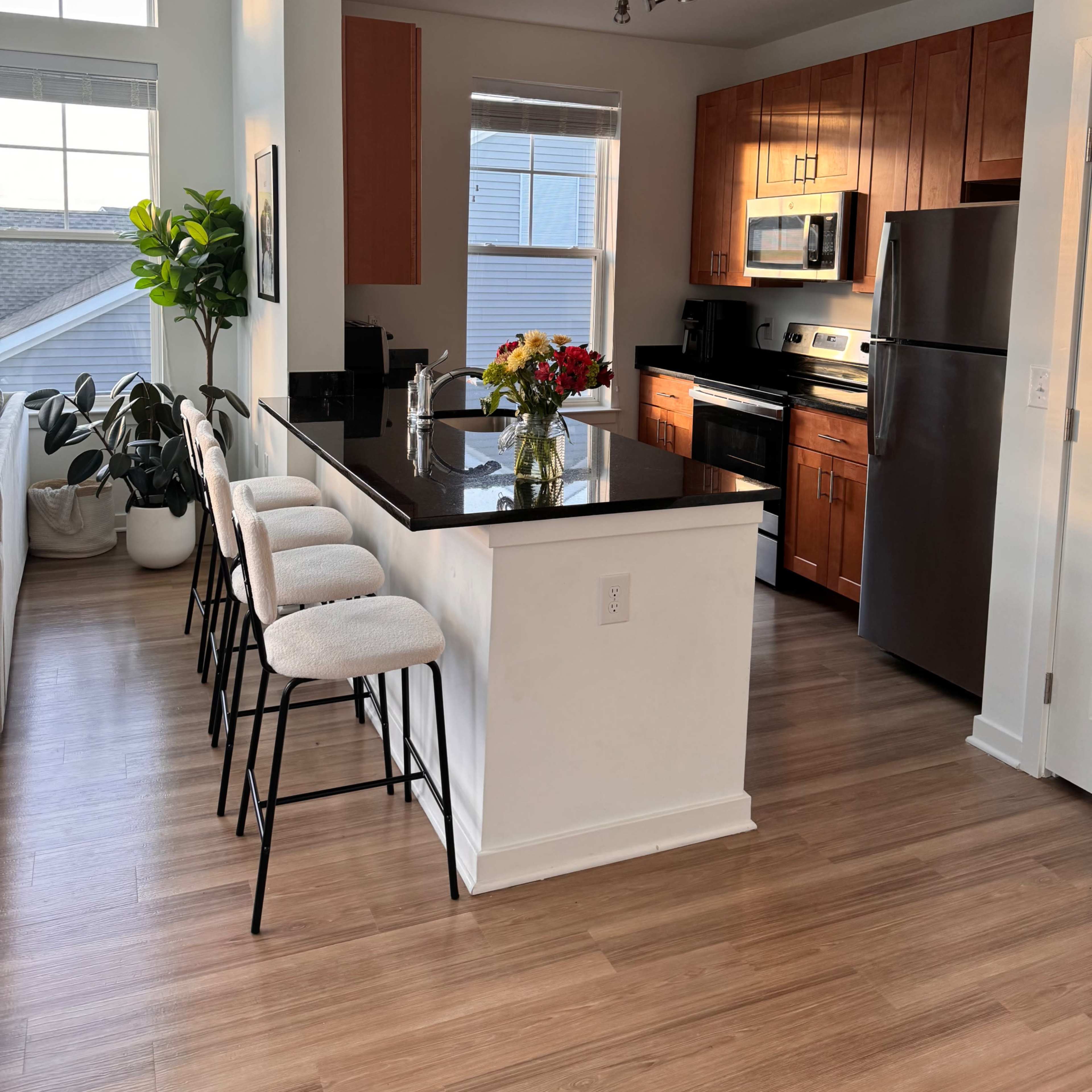 A modern kitchen with wooden cabinets, a black countertop, and a vase of flowers on the island, complemented by bar stools and large windows letting in natural light.