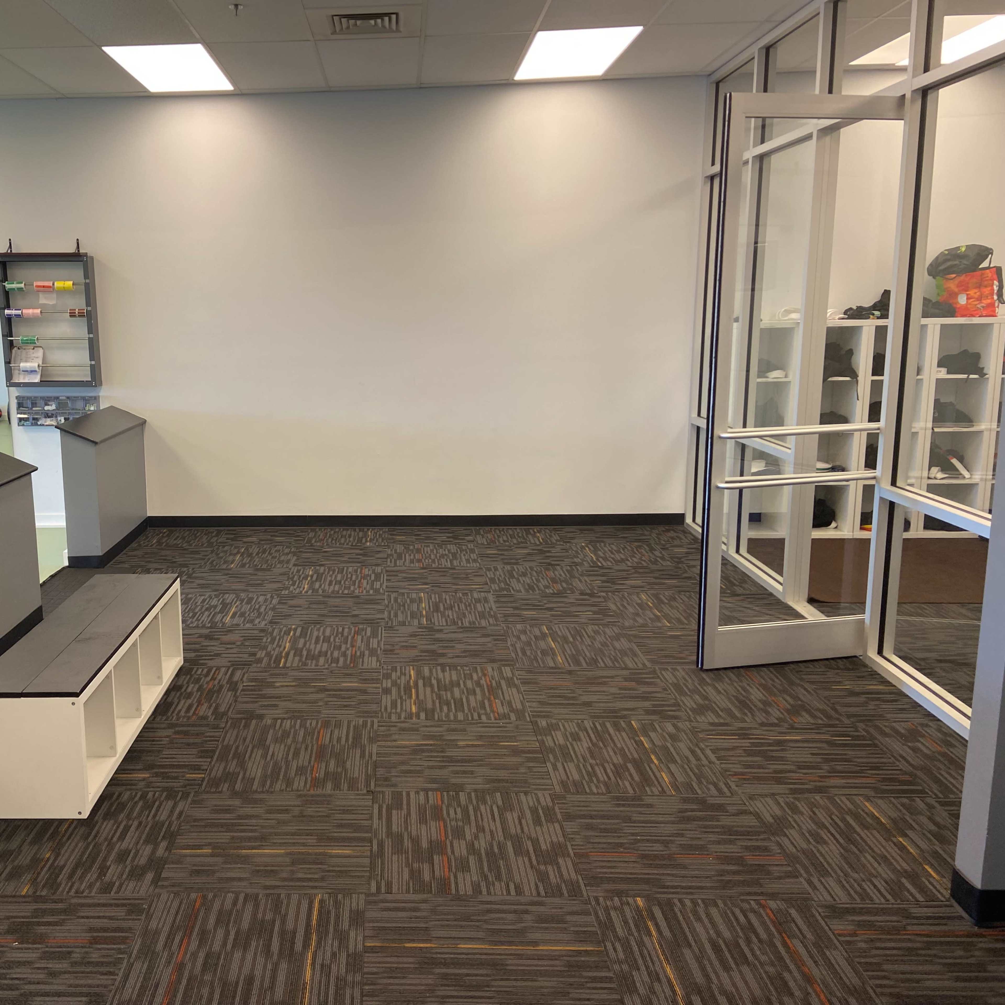 A spacious area with a gray and black carpet, a low white bench, and large windows leading to an outside entrance, alongside a wall with fitness equipment on display.