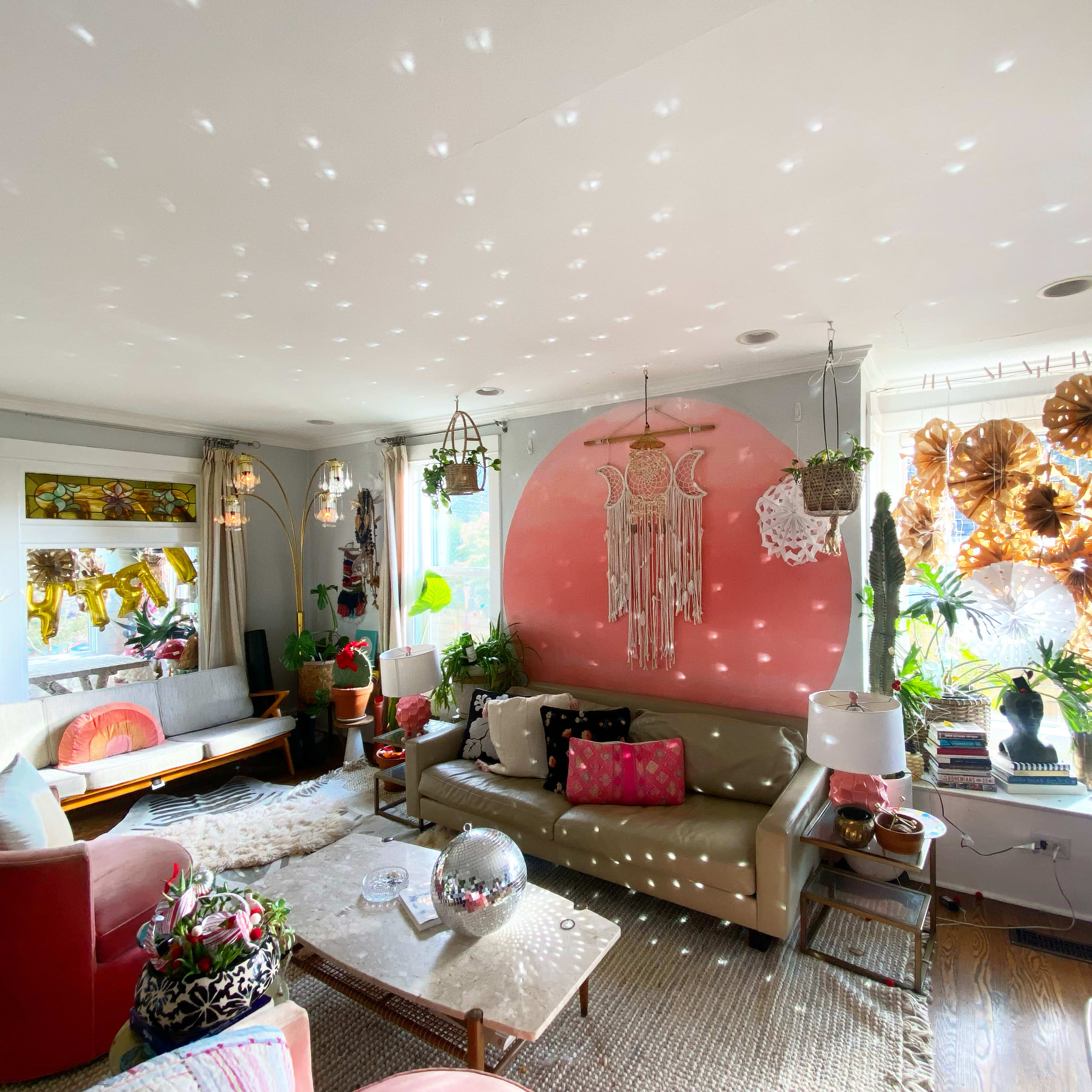 The living room features a mix of modern furniture and decorative plants, with a large pink circular wall art piece and colorful light reflections on the ceiling.