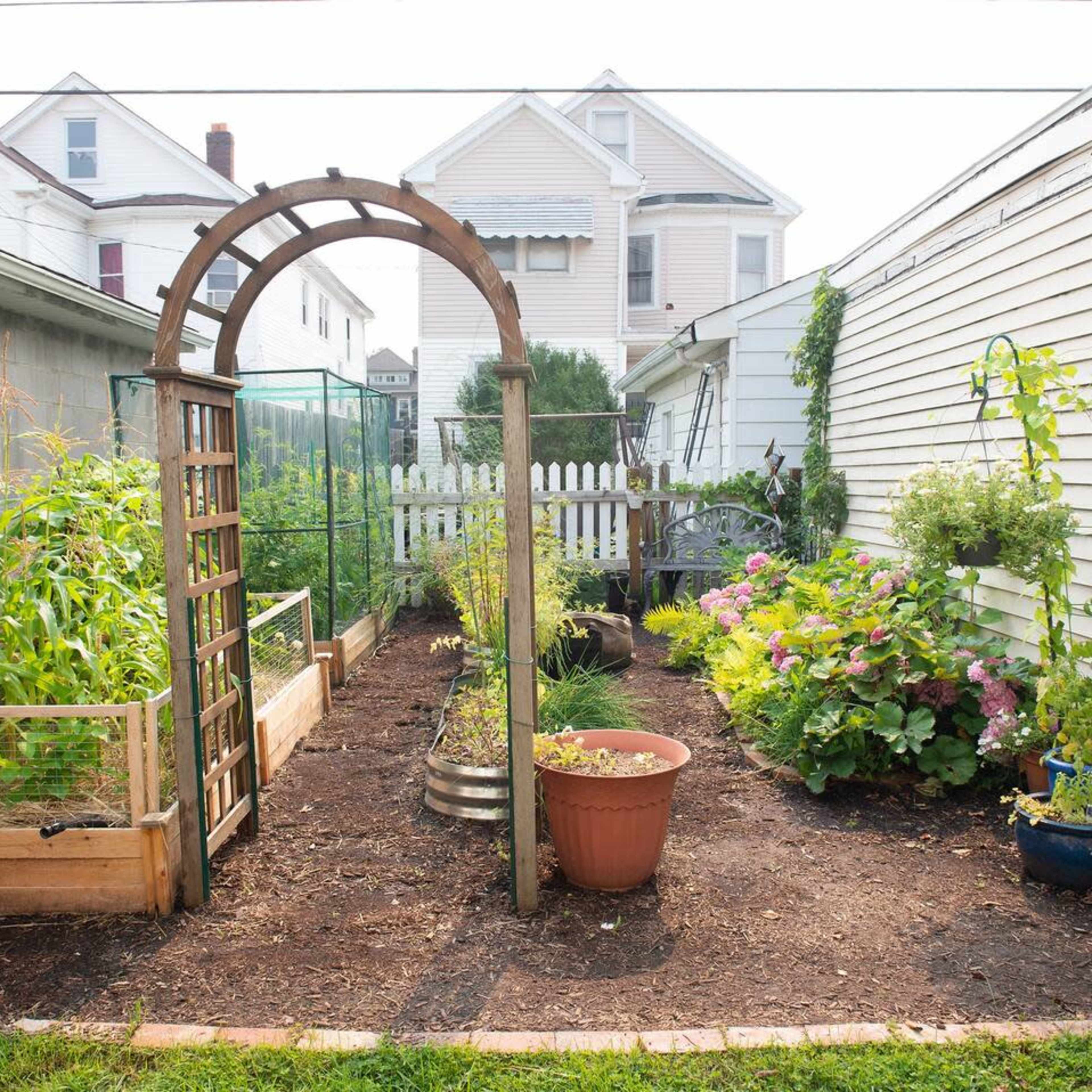 A garden is enclosed by wooden archways and features raised beds, potted plants, and a variety of greenery along a narrow pathway.