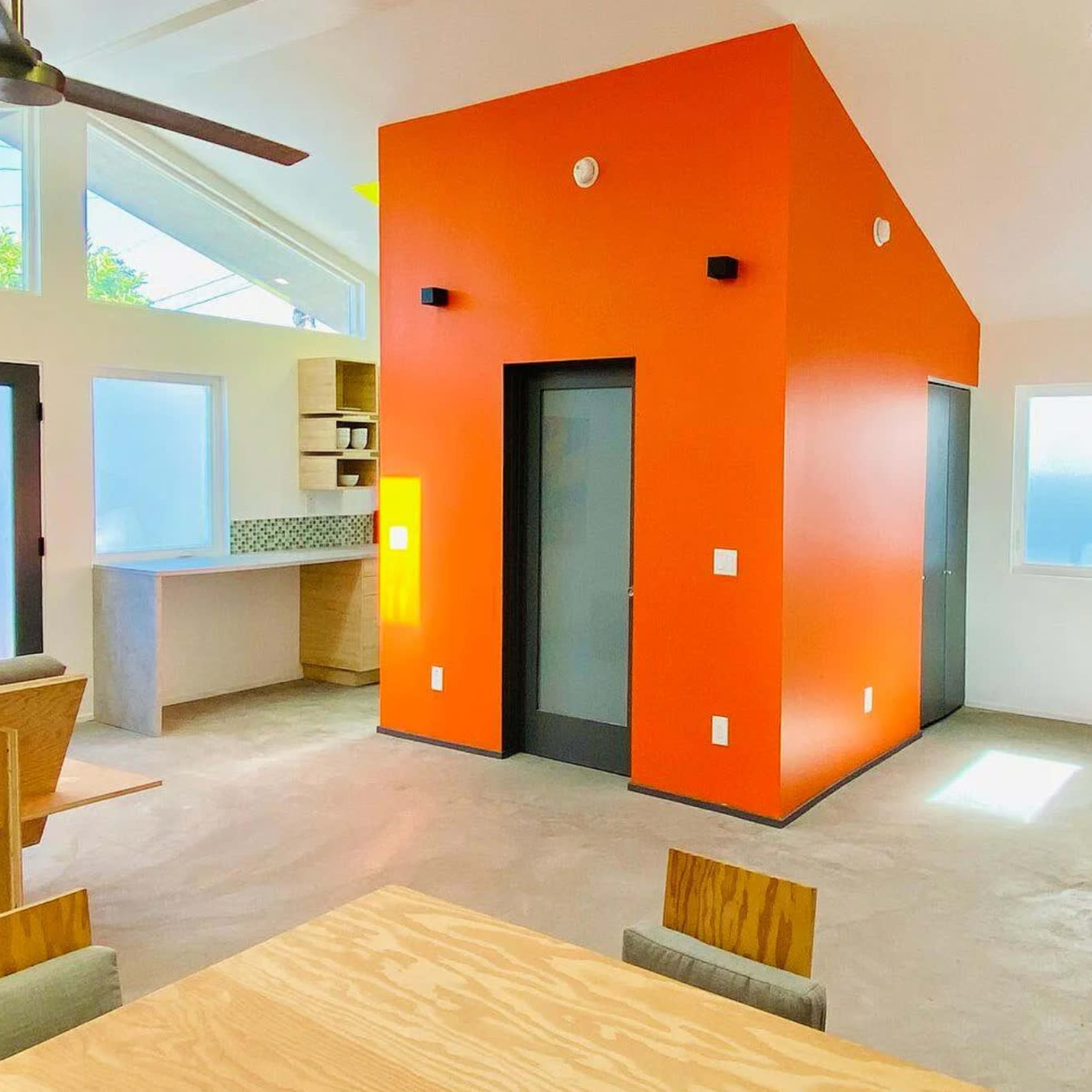A modern, open-concept living space featuring a striking orange wall, concrete flooring, and large windows with a view.