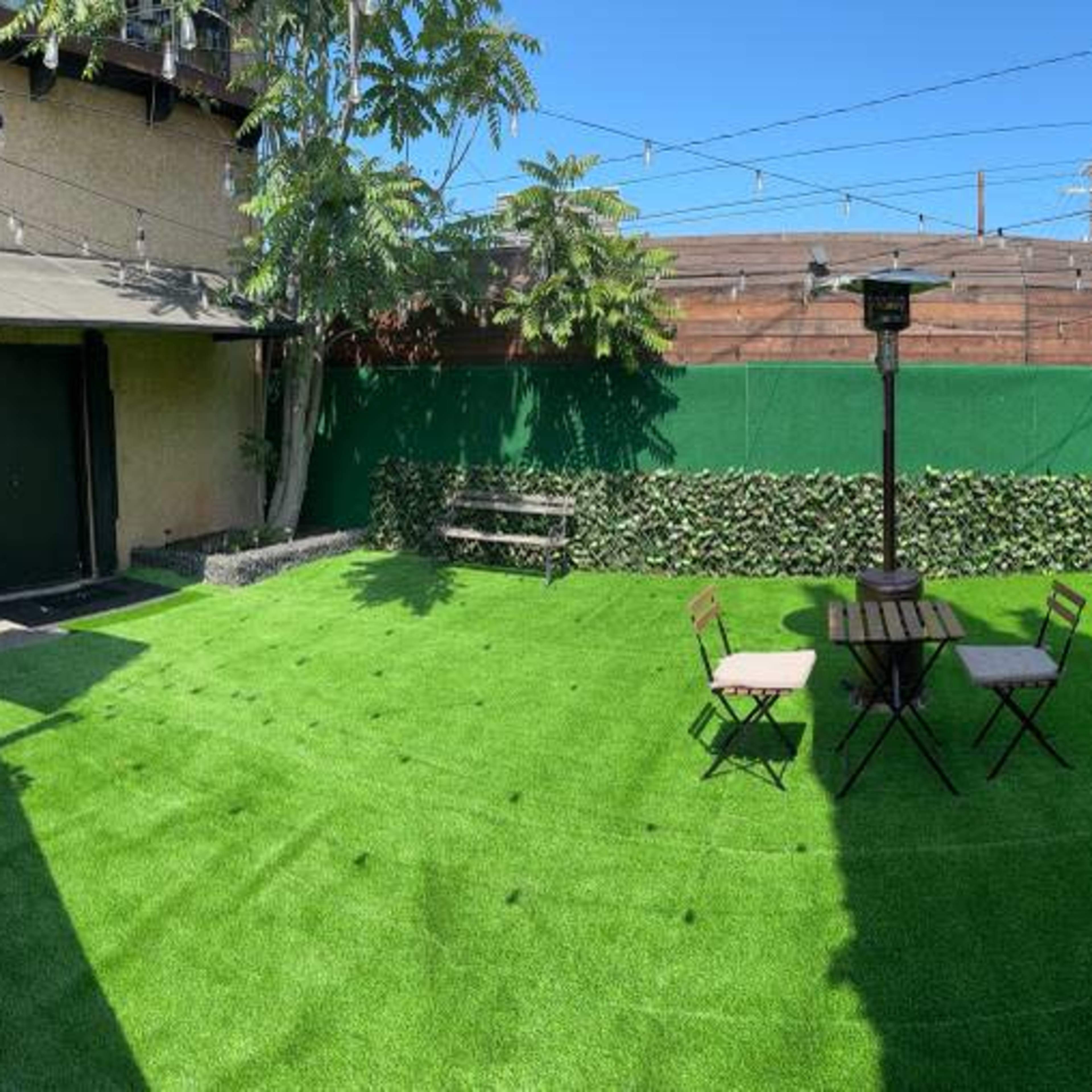 A panoramic view of a green artificial grass yard featuring a table with four chairs, surrounded by hedges and a tall heater.