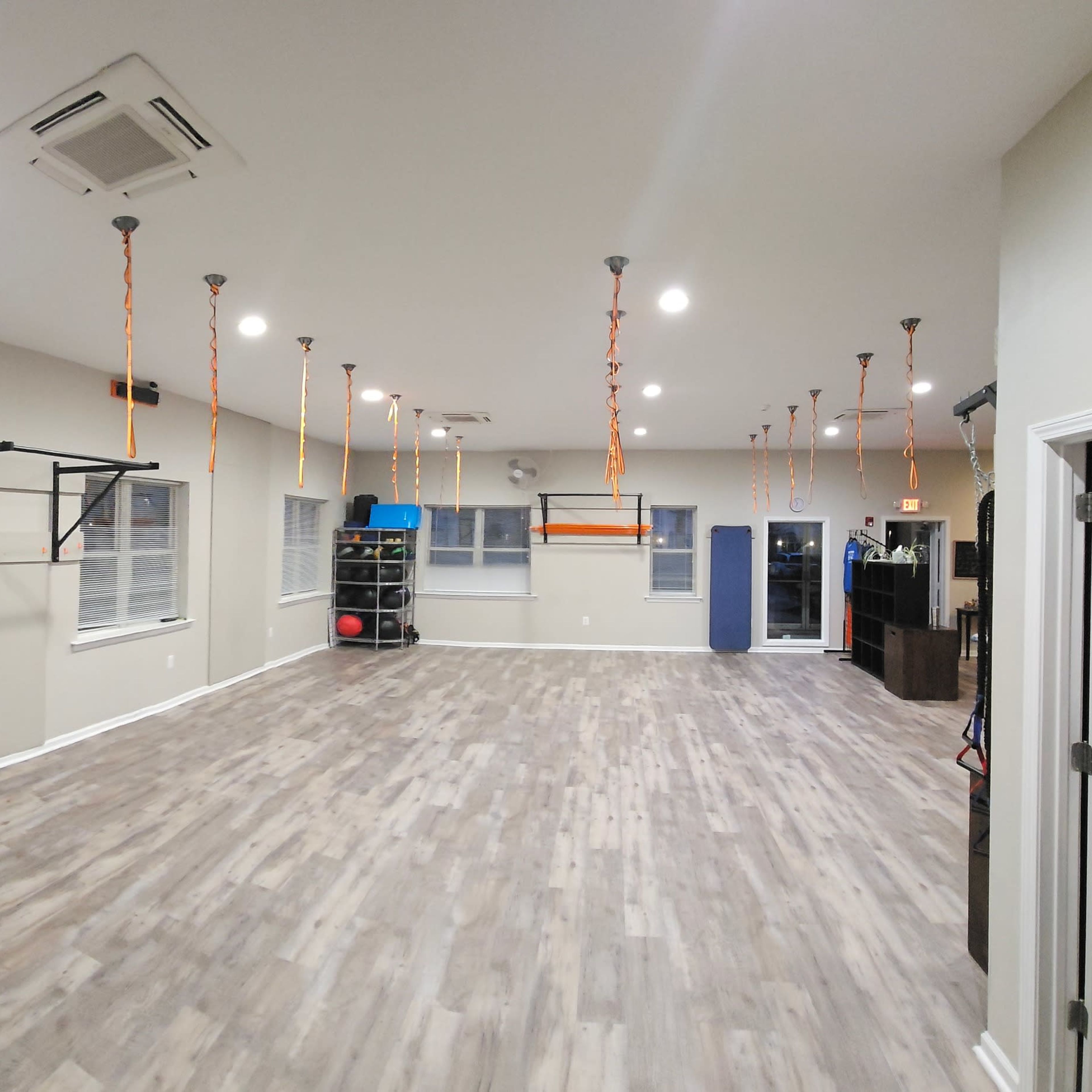 A spacious, well-lit gym with wooden flooring, various exercise equipment along the walls, and suspended ropes hanging from the ceiling.