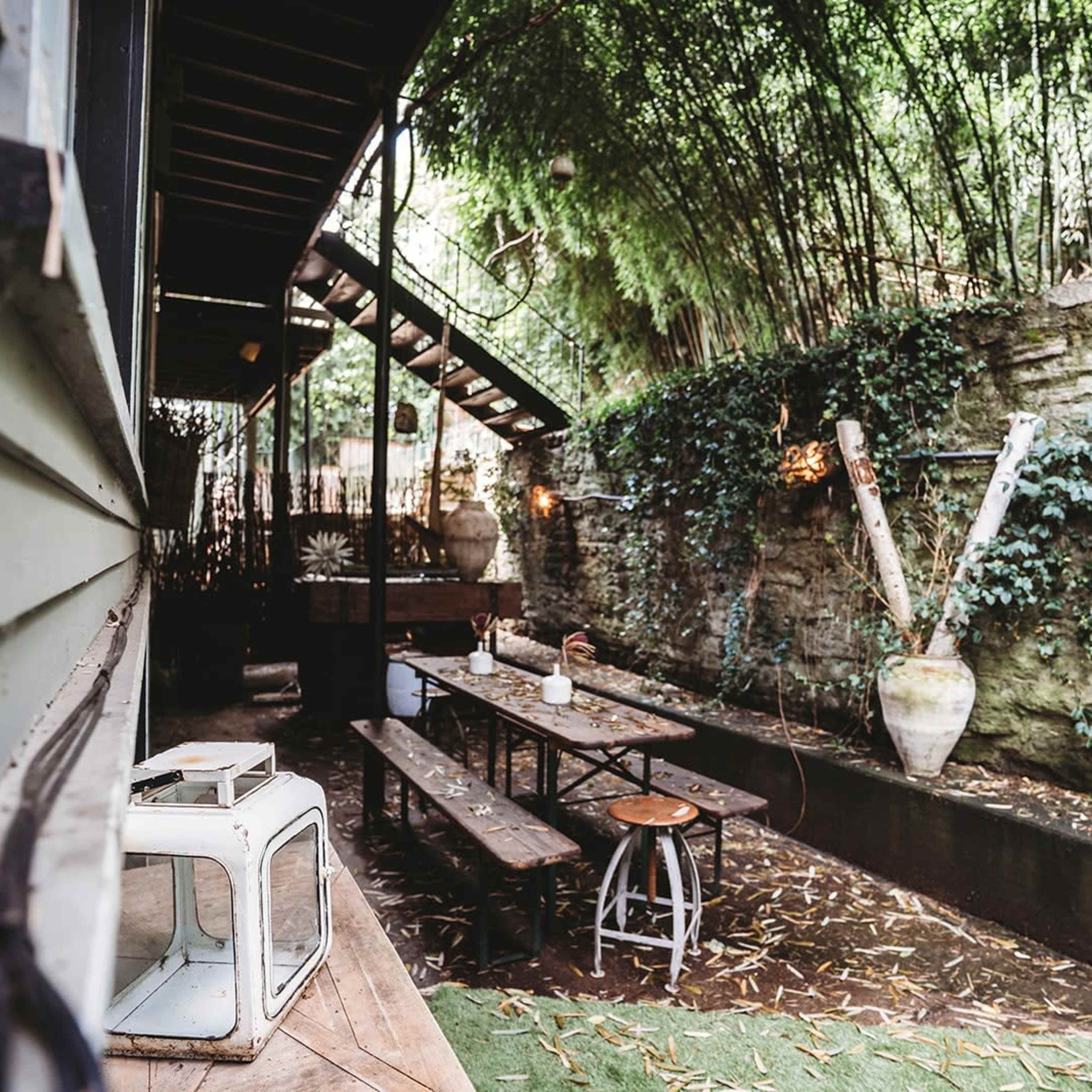 A small outdoor seating area features wooden benches and tables, surrounded by greenery and stone walls.