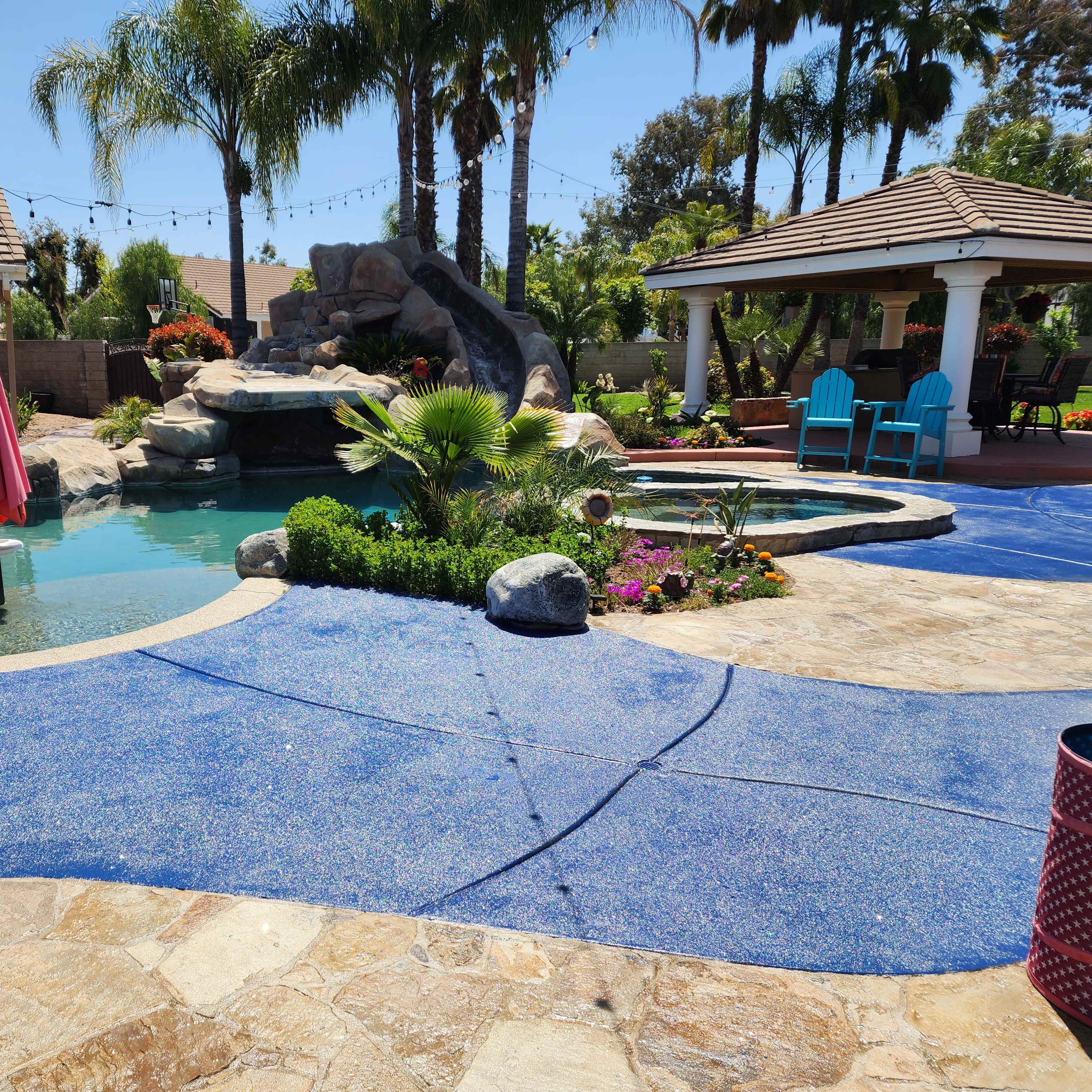 A backyard pool area featuring a rock waterfall, tropical landscaping, and a cabana with lounge chairs.