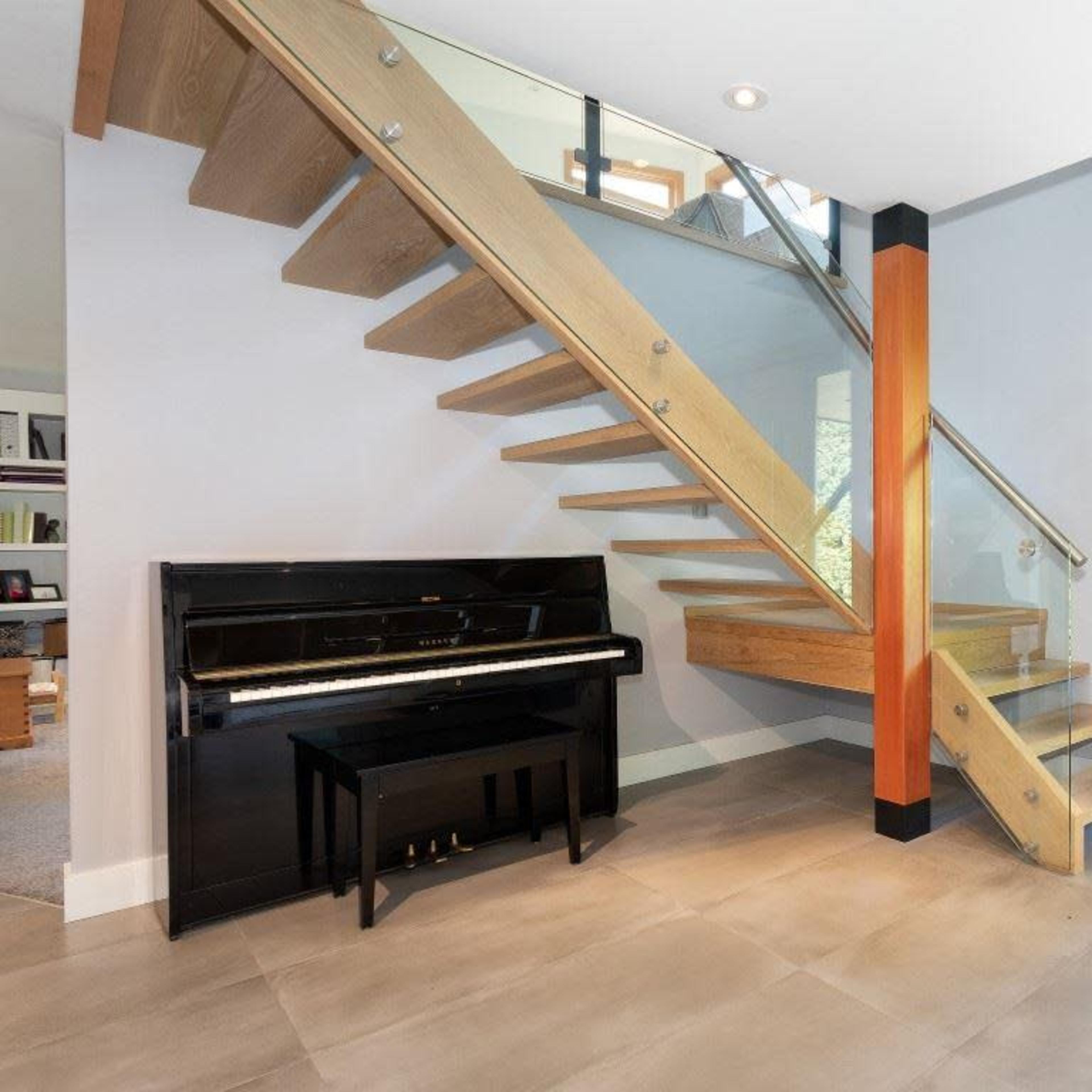 A modern entryway featuring a wooden staircase with glass railings and a black piano against a wall next to a large wooden door.