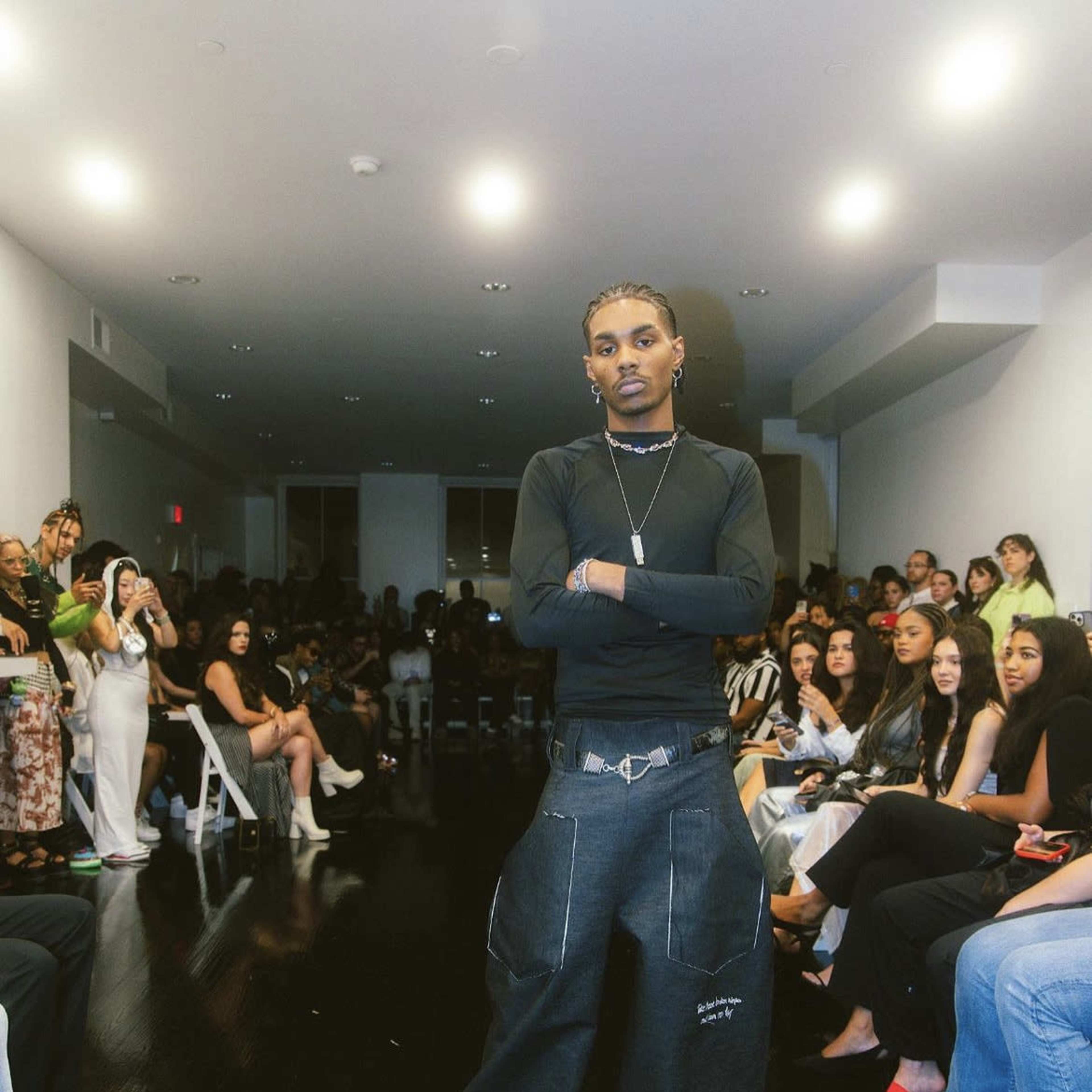 A model poses in a dimly lit room filled with seated attendees during a fashion show.