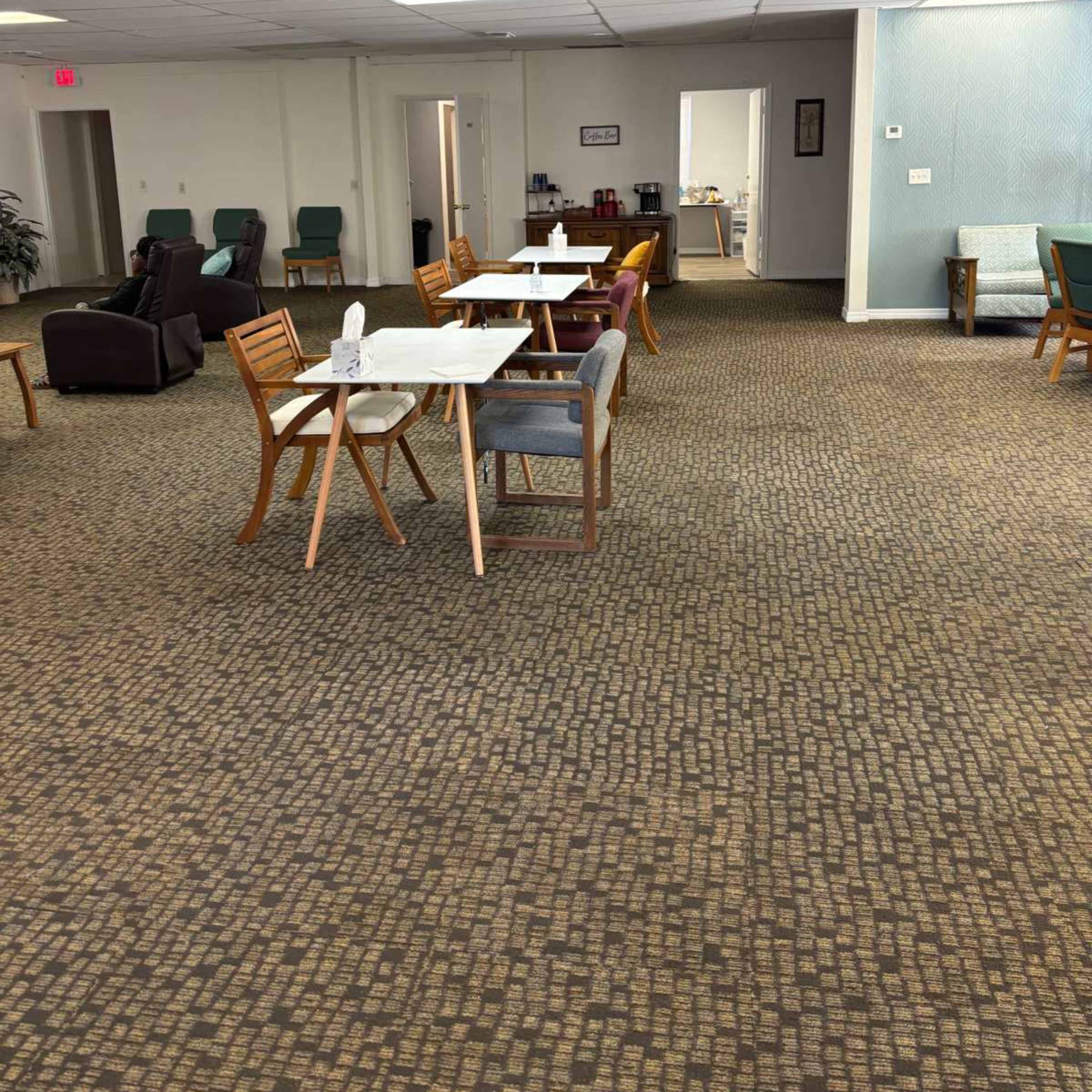 A spacious room with several tables and chairs arranged on a patterned carpet, leading toward a doorway in the background.