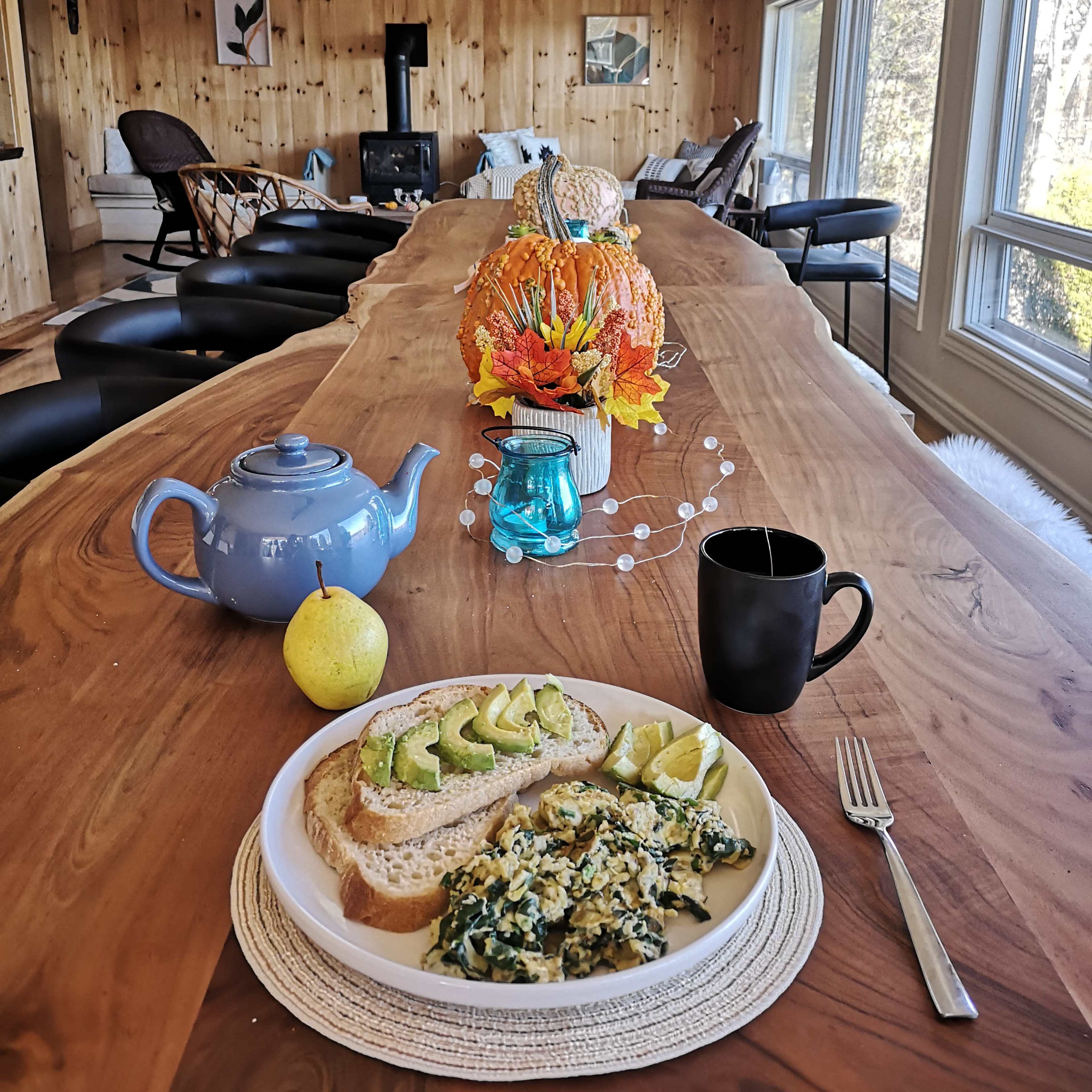 A long wooden dining table is set with a plate of scrambled eggs and avocado on toast, a pear, a black mug, and a blue teapot, with a decorative centerpiece of autumn flowers.