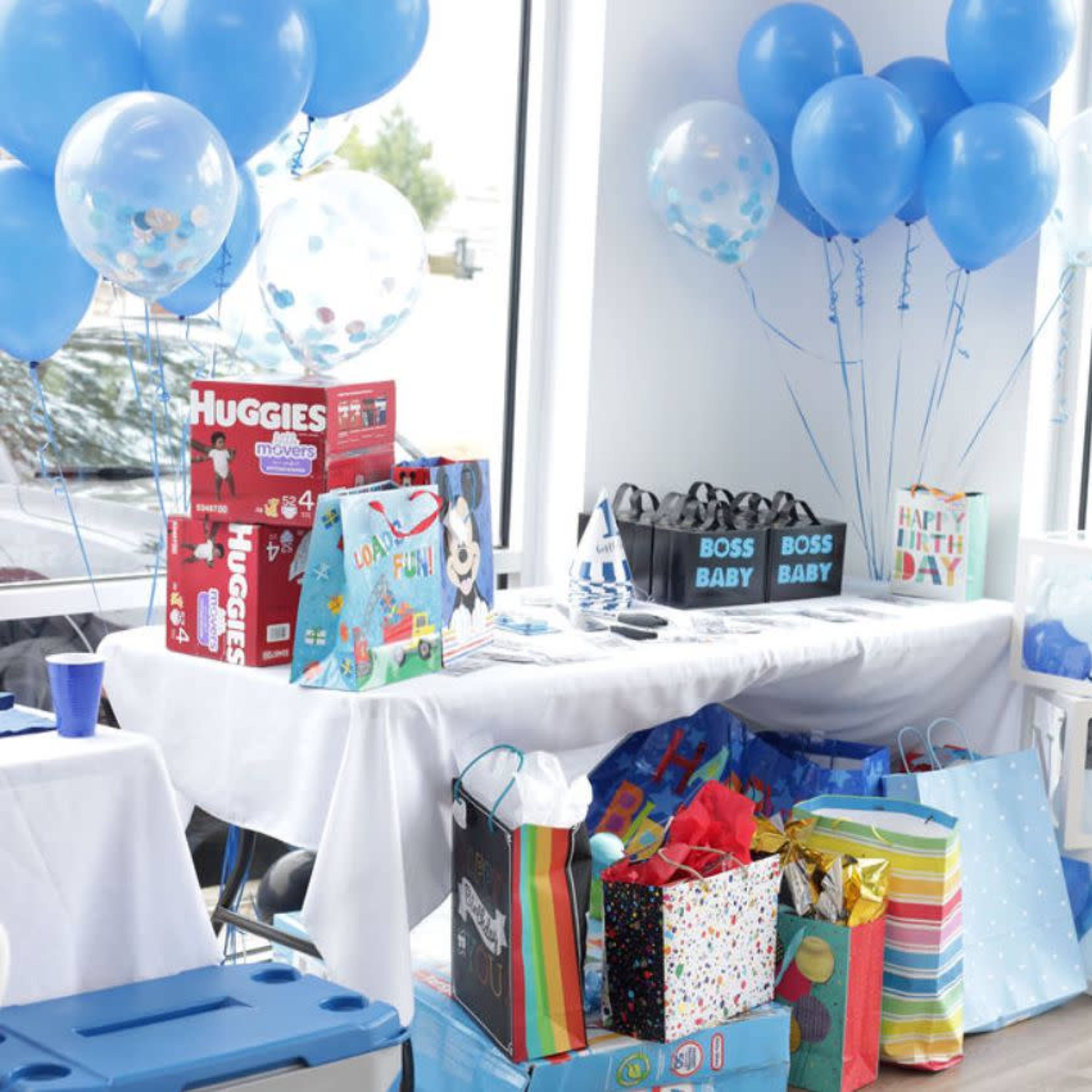 The scene shows a table decorated for a birthday party, featuring colorful gift bags, wrapped presents, and blue balloons.