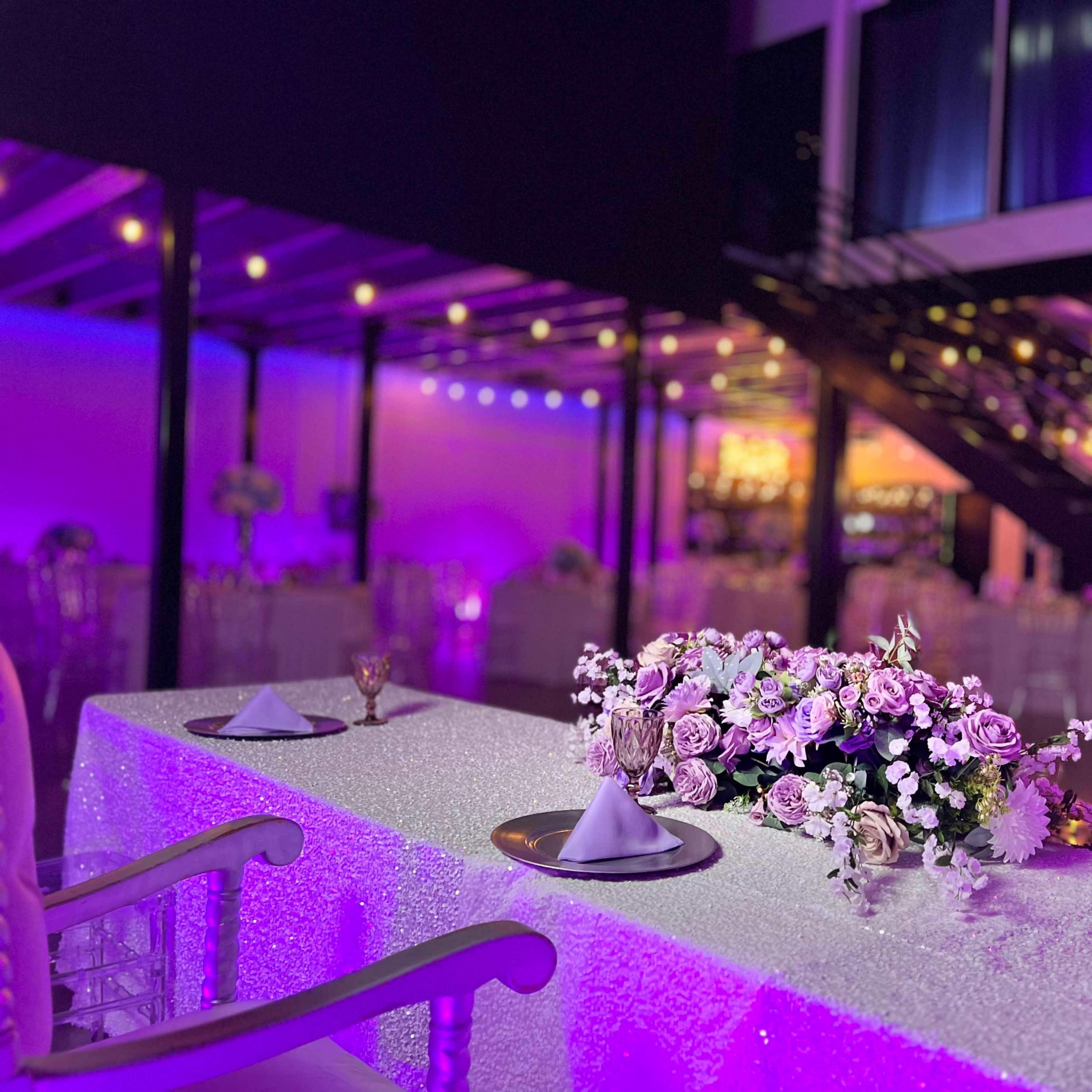 A decorated table with a floral centerpiece is set up in a brightly lit event space featuring rows of tables and a staircase in the background.