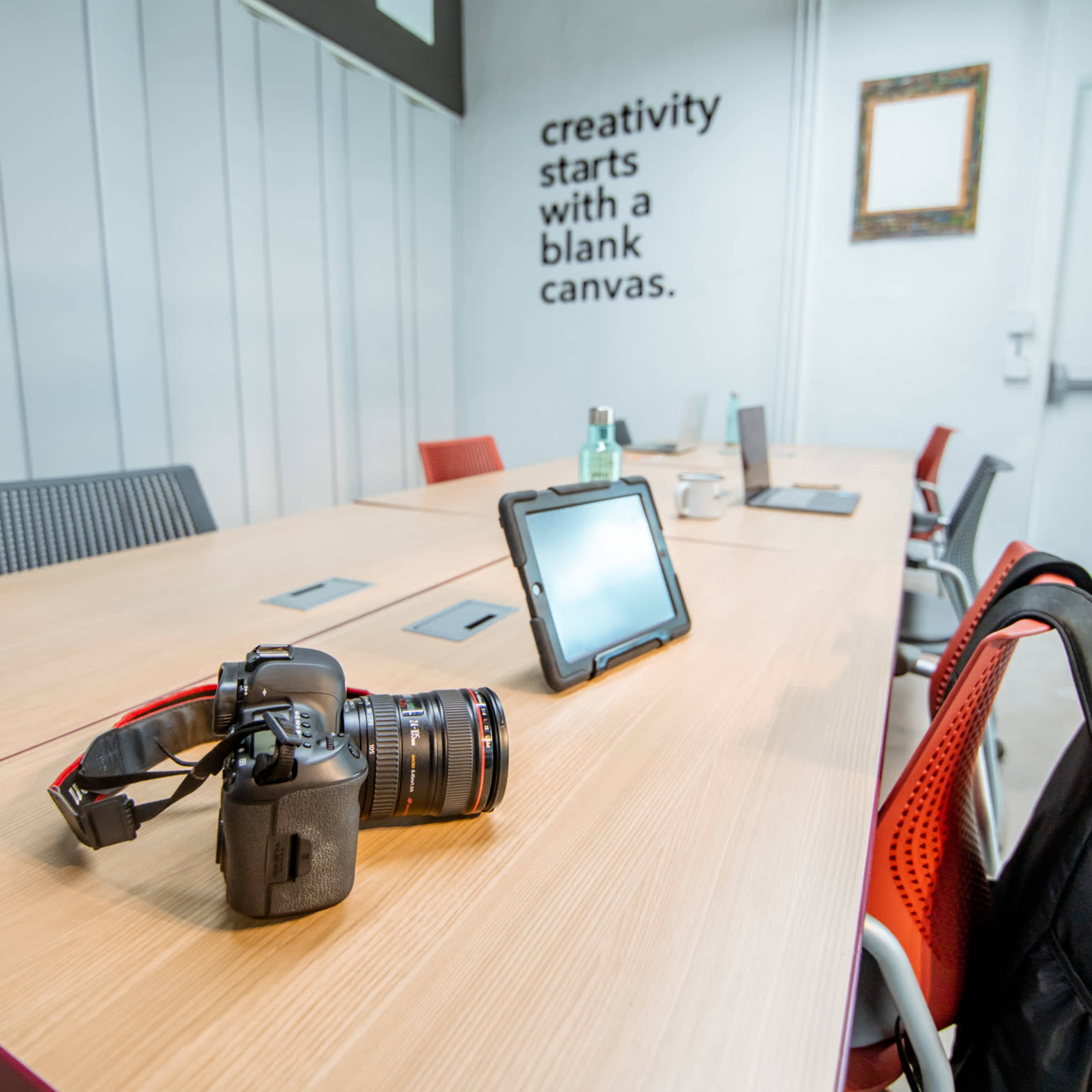 A conference room with a large wooden table, equipped with a camera, tablet, and several chairs, featuring a wall with an inspirational quote about creativity.
