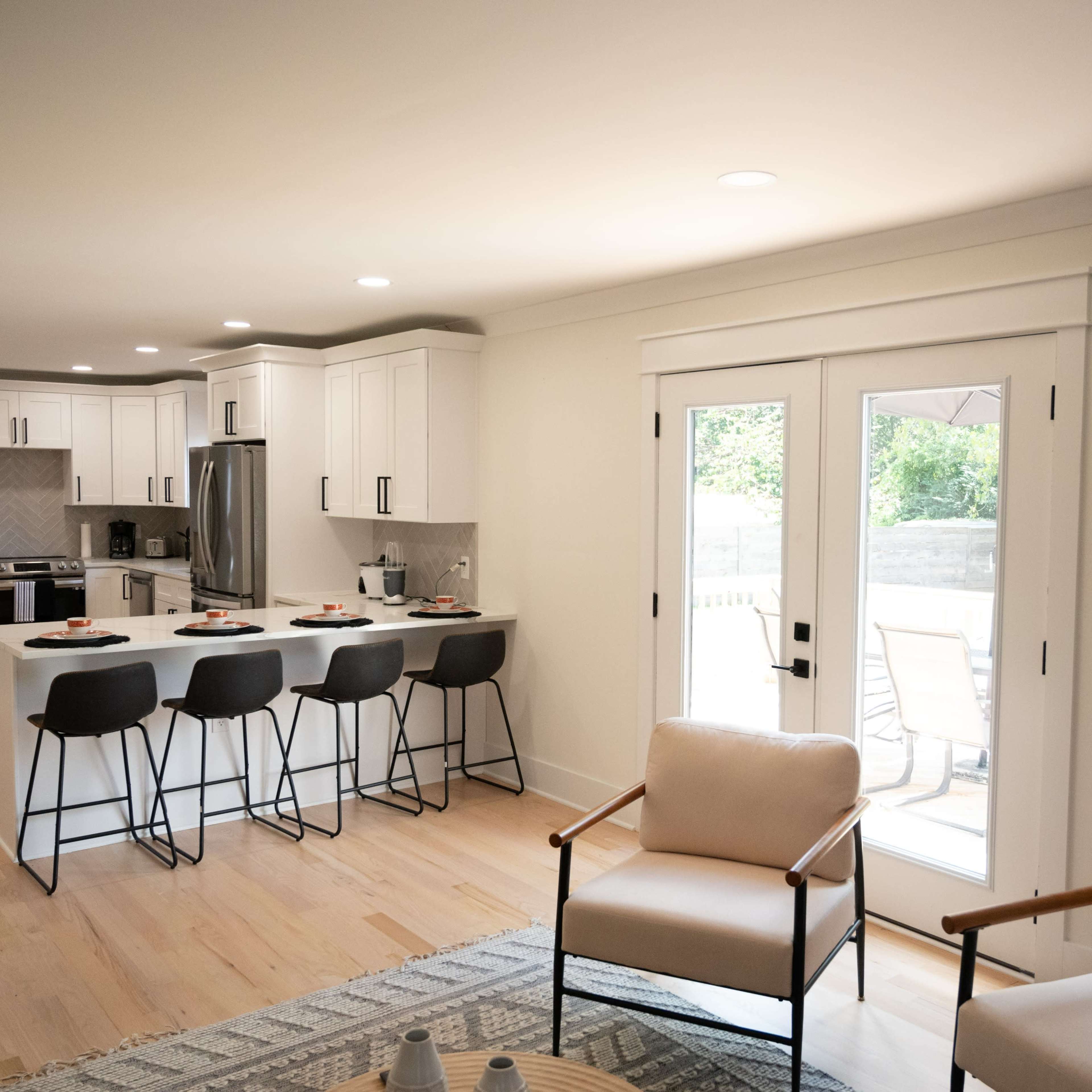 A modern kitchen with white cabinetry and a central island adjoins a living area with two armchairs and sliding glass doors leading to an outdoor patio.