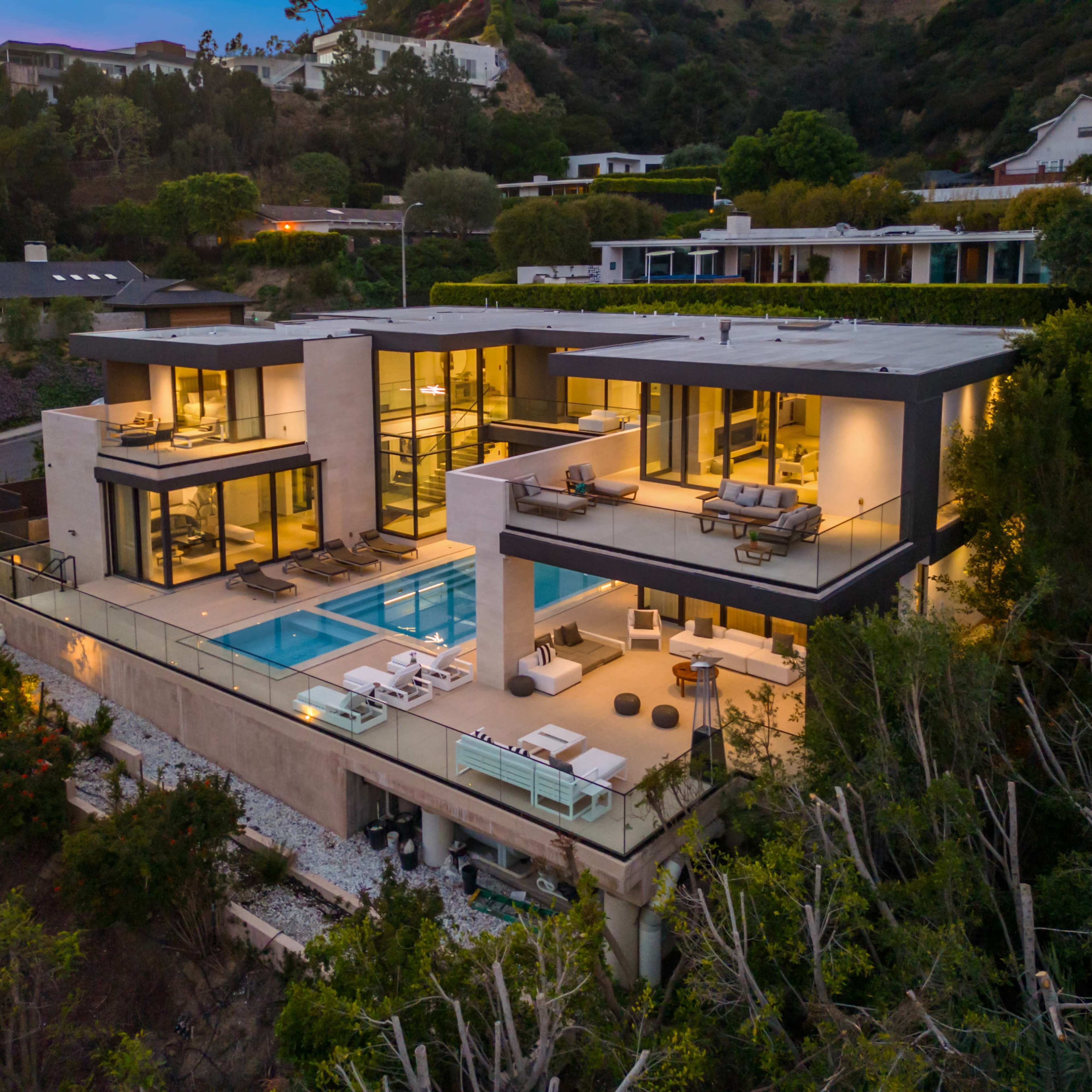 A modern, multi-level house with large glass windows and a swimming pool is situated on a hillside surrounded by greenery.