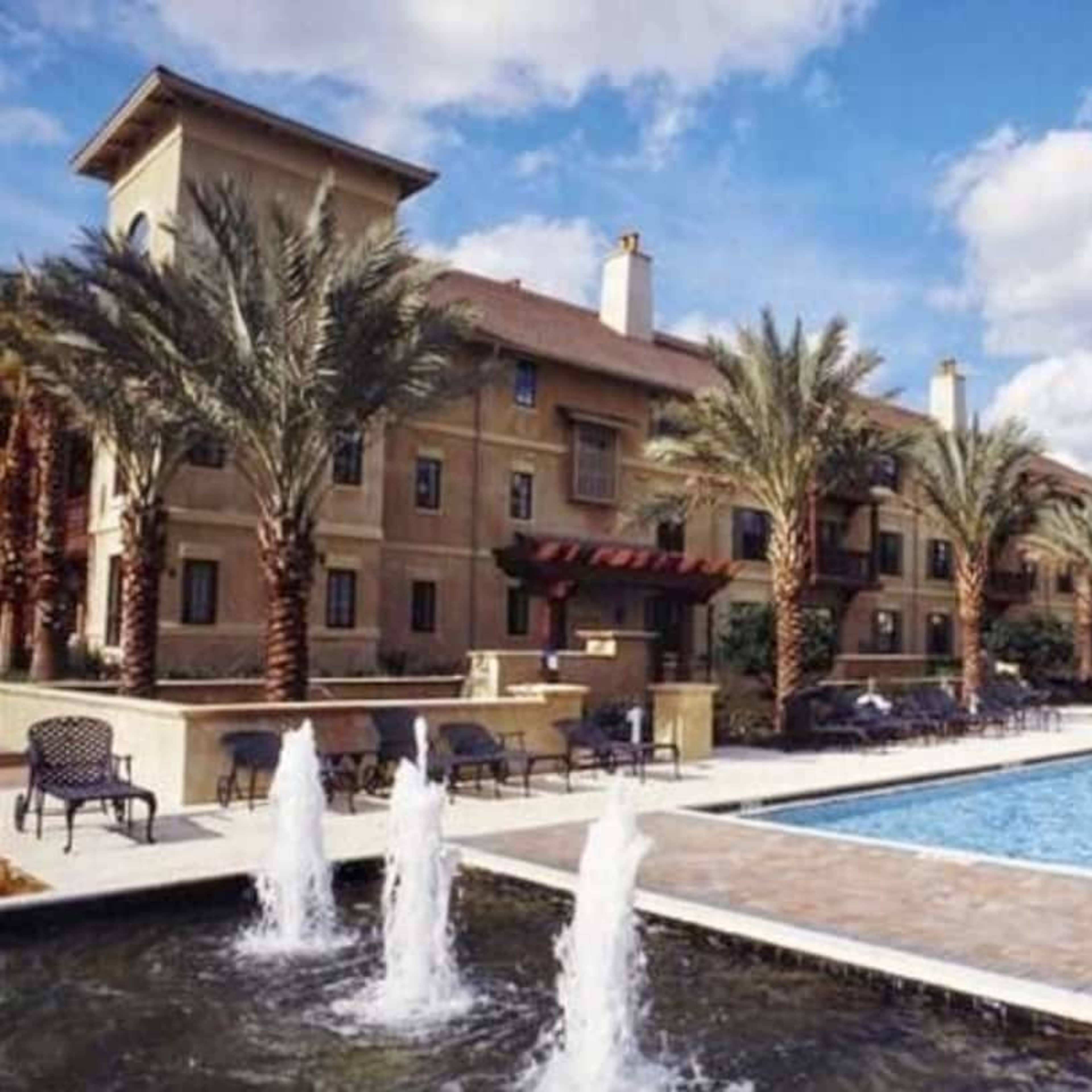 An apartment complex with a swimming pool, palm trees, and fountains in the foreground.