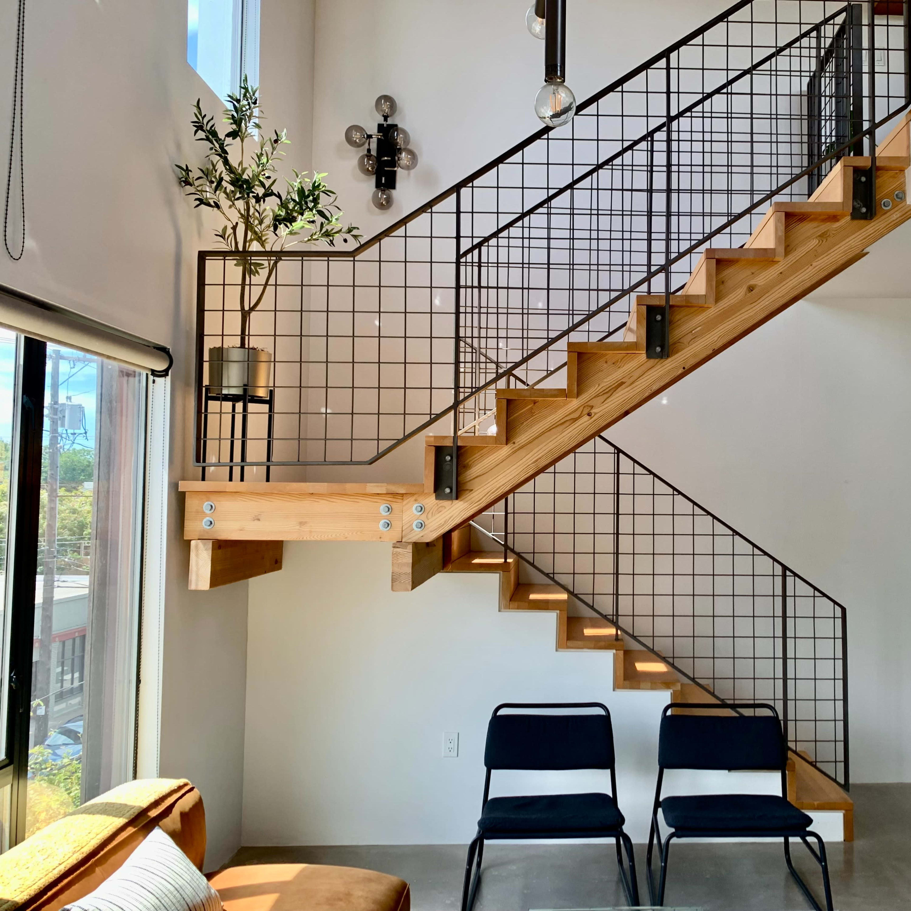 A modern staircase made of wood with a metal railing, situated next to large windows and two black chairs on a concrete floor.