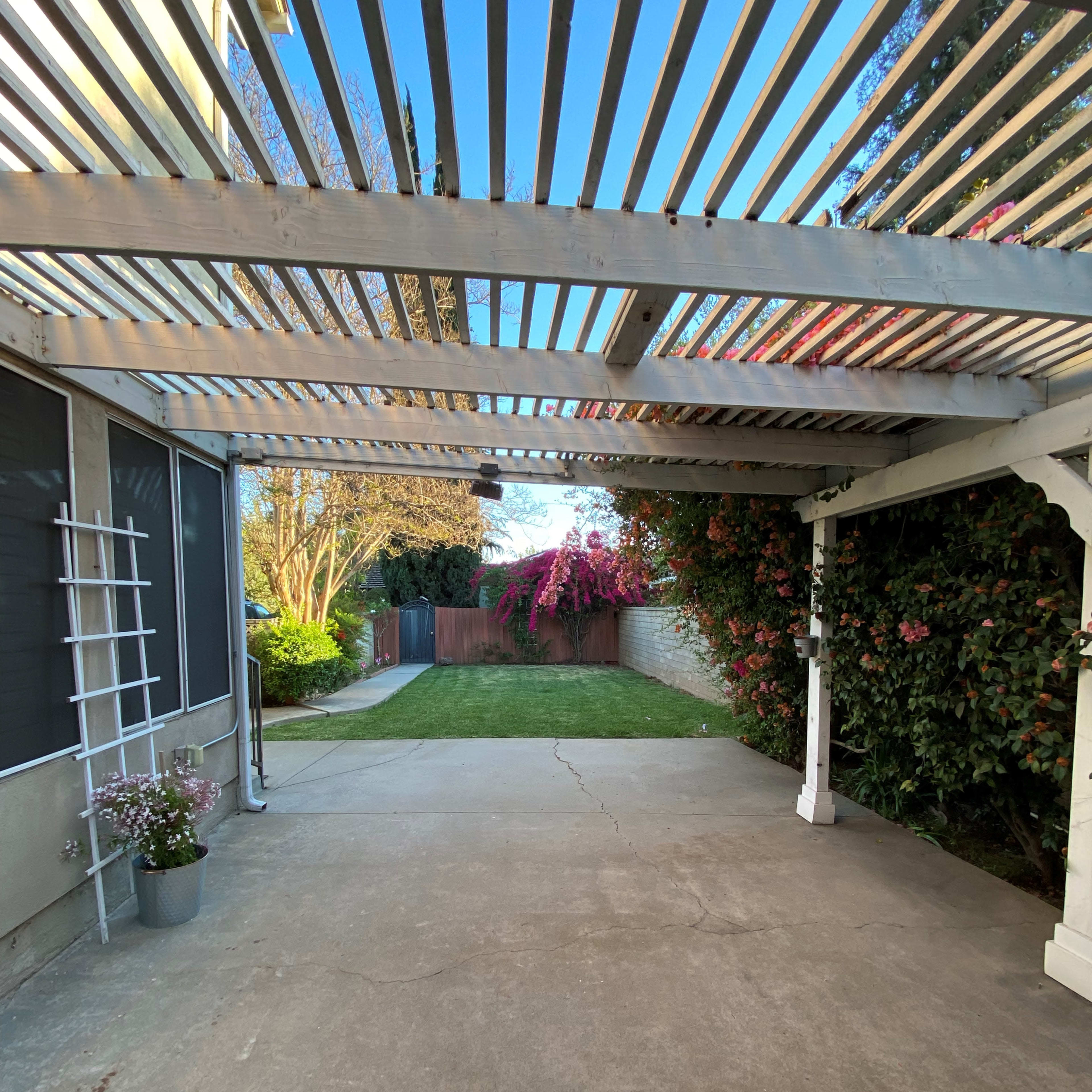 A patio area with a slatted overhead structure, concrete flooring, and green grass beyond, bordered by flowering plants and a wooden fence.