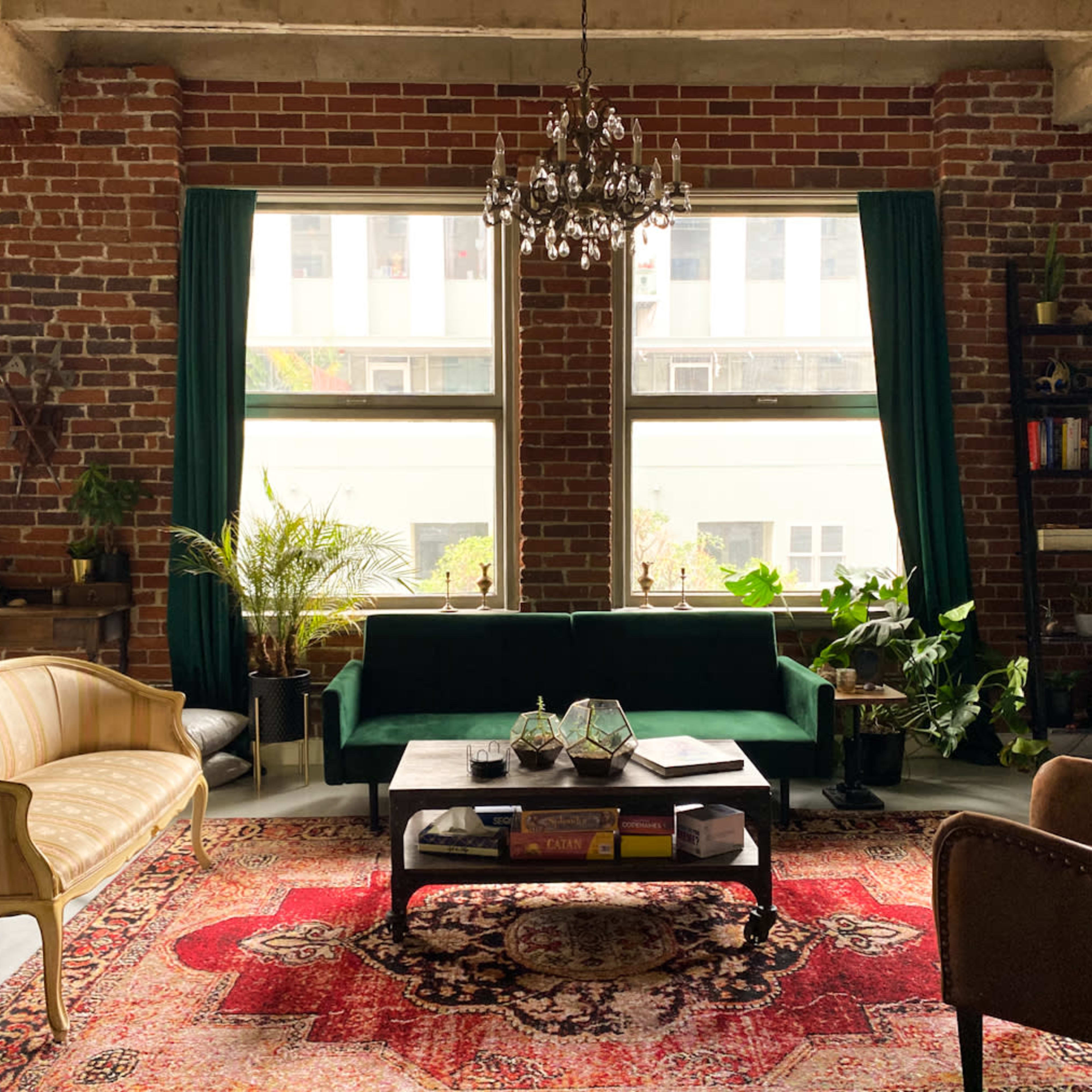 A cozy living room features a green sofa, a patterned rug, and large windows surrounded by brick walls and indoor plants.
