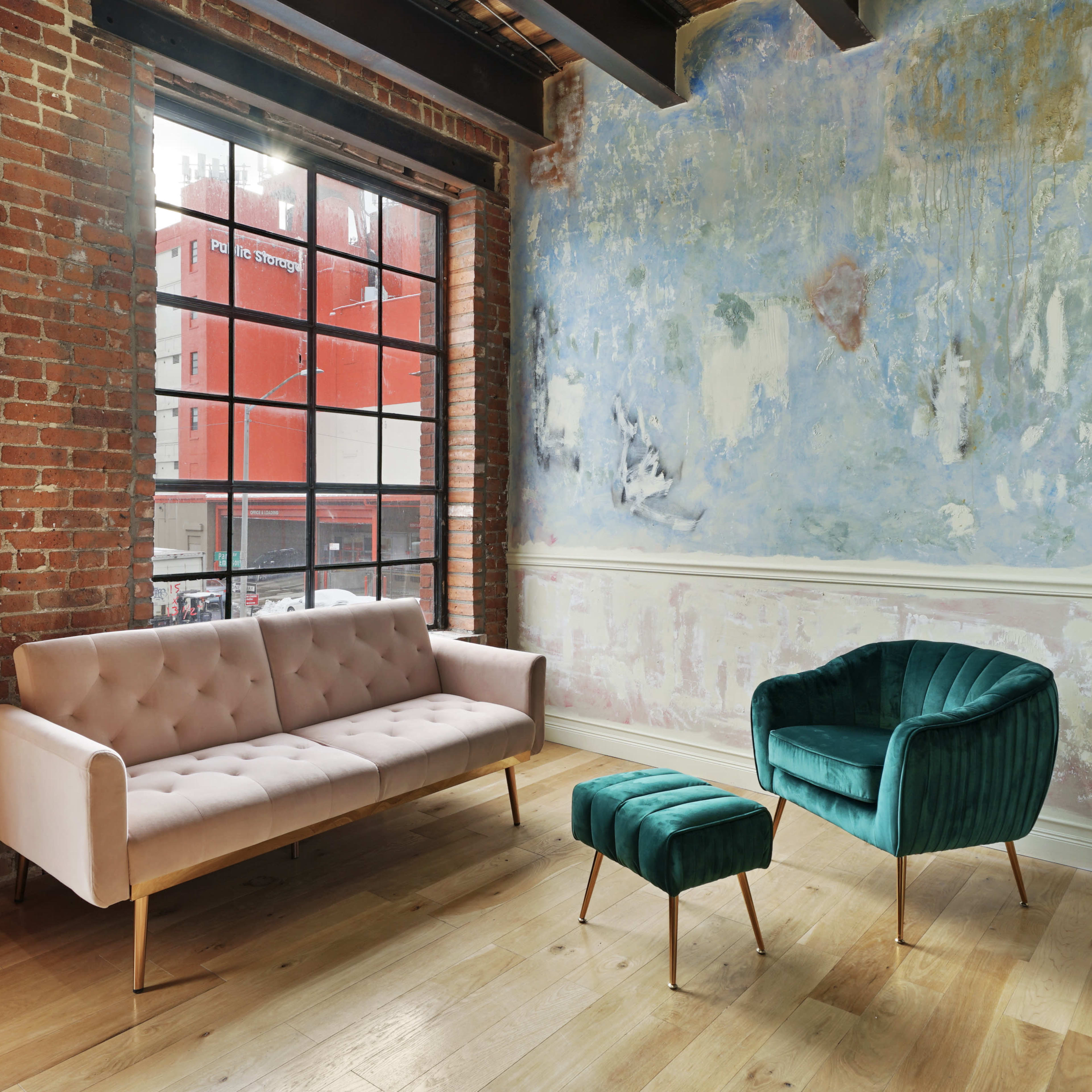 A living space with a pink tufted sofa, a green velvet armchair, and a matching ottoman, set against a backdrop of exposed brick walls and a painted blue wall with large windows.