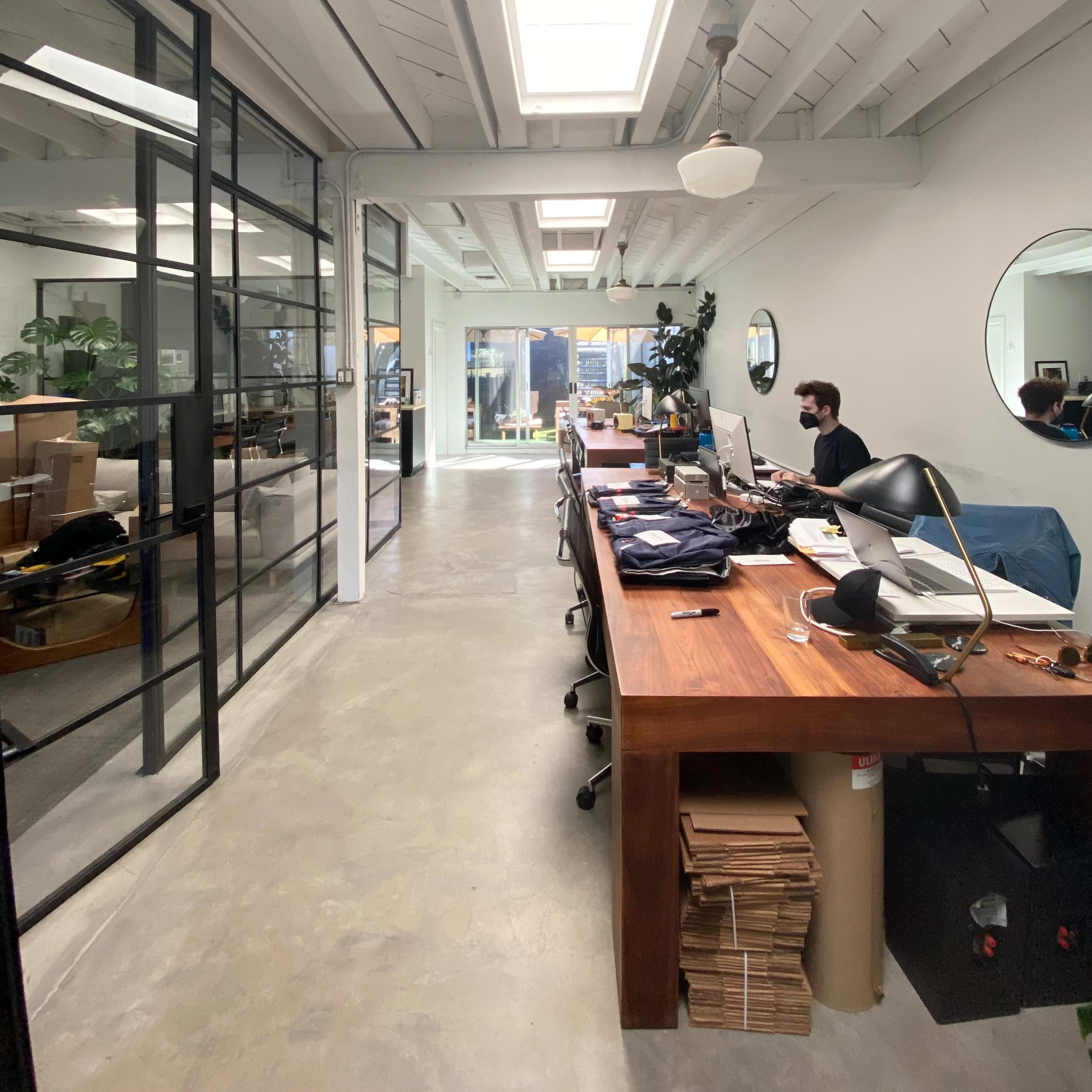 A spacious office area features a long wooden table with scattered items, large windows allowing natural light, and a visible workspace with a man seated at a desk.