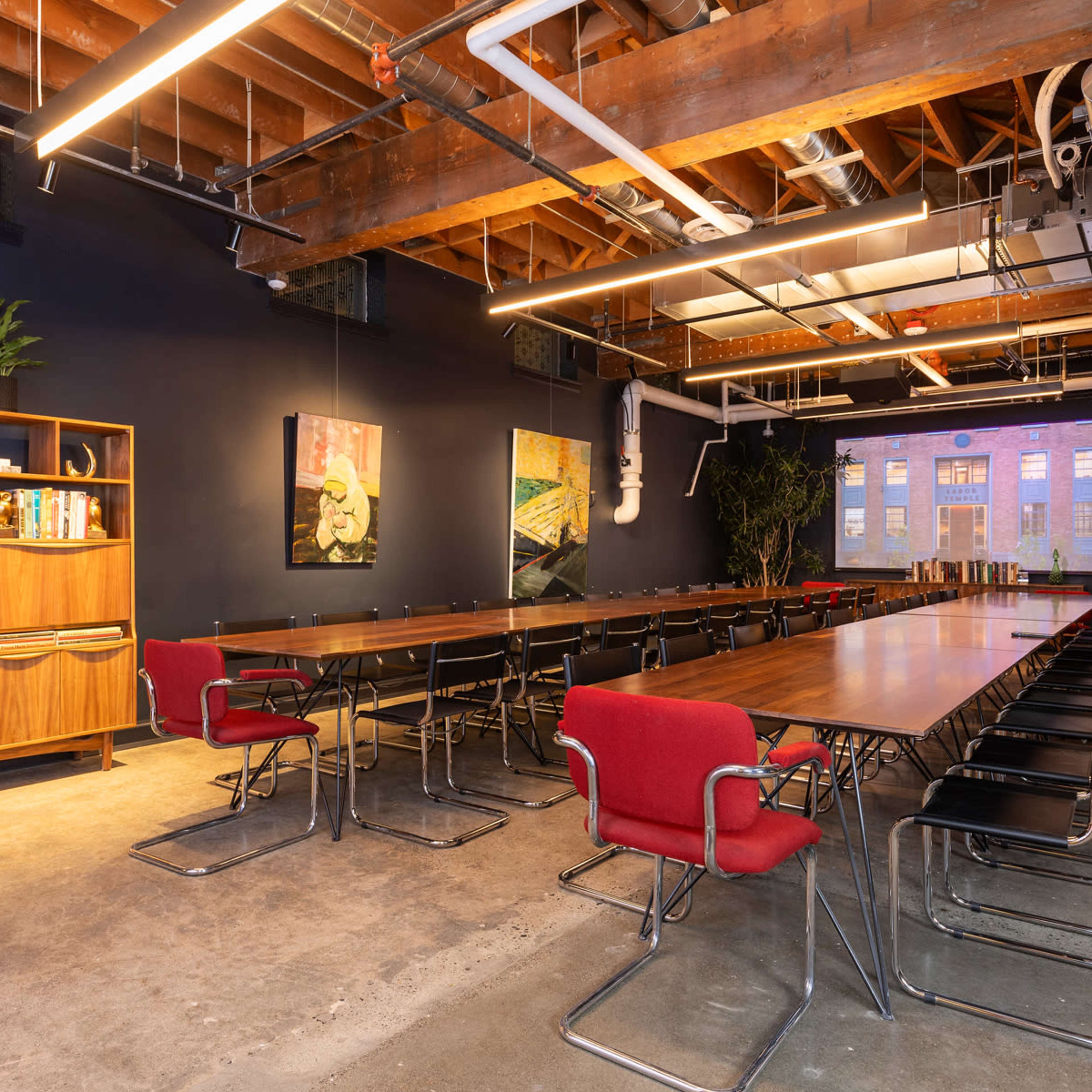A conference room features long wooden tables with red chairs, a wooden shelving unit, and large windows displaying an outdoor view.