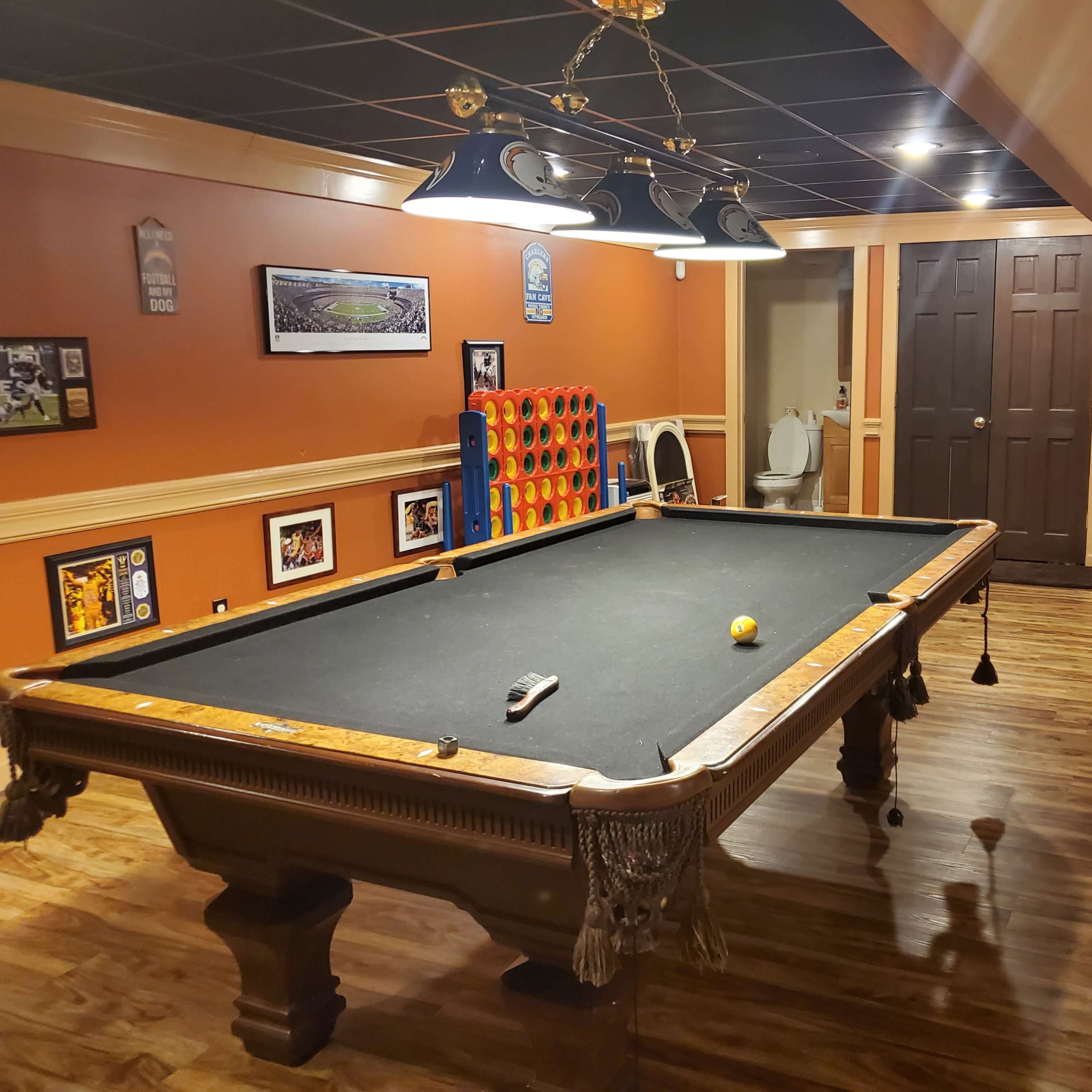 A recreation room featuring a pool table, wooden flooring, orange walls, framed pictures, and a bathroom door in the background.