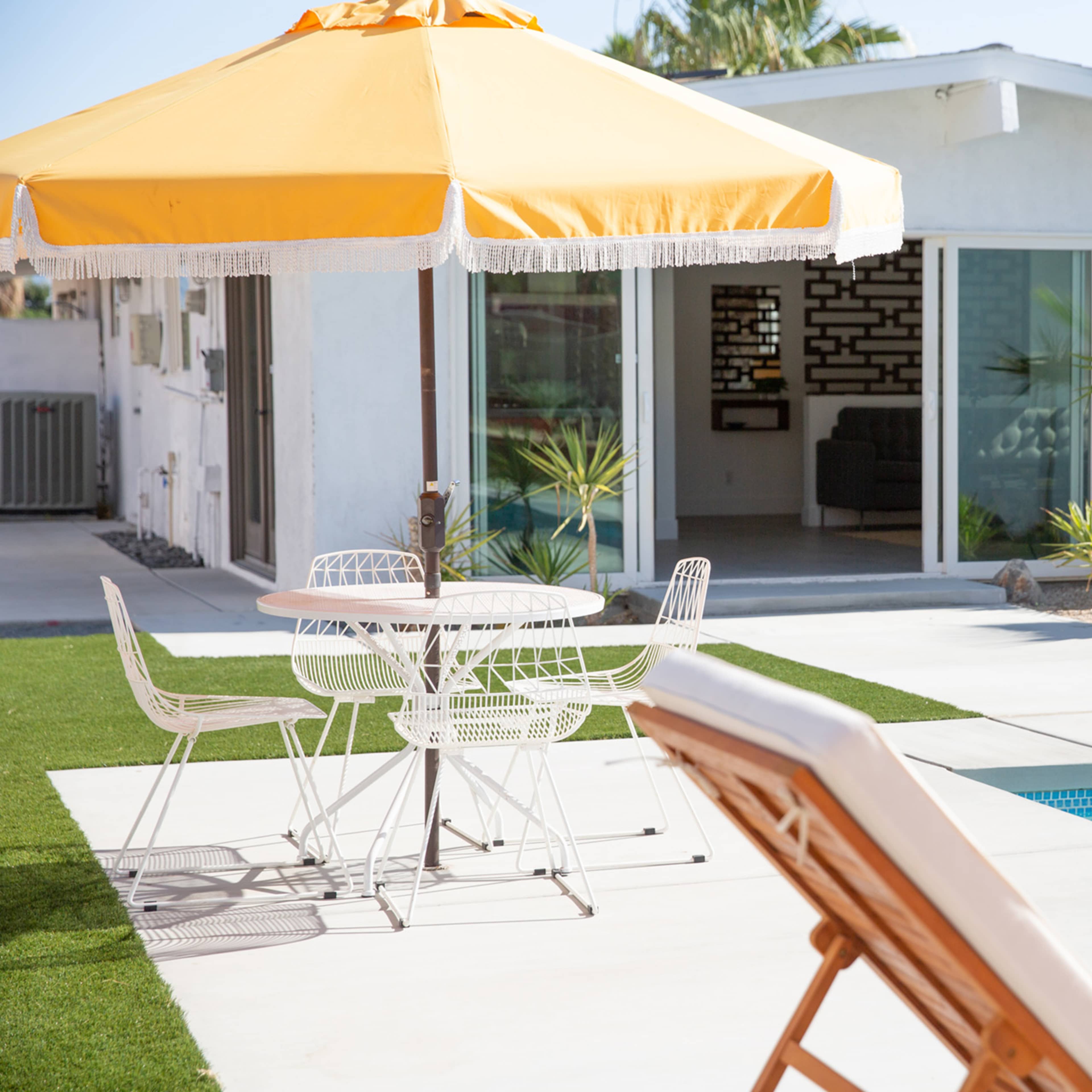 A yellow umbrella shades a white patio table and chairs near a swimming pool, with a modern home and palm trees in the background.