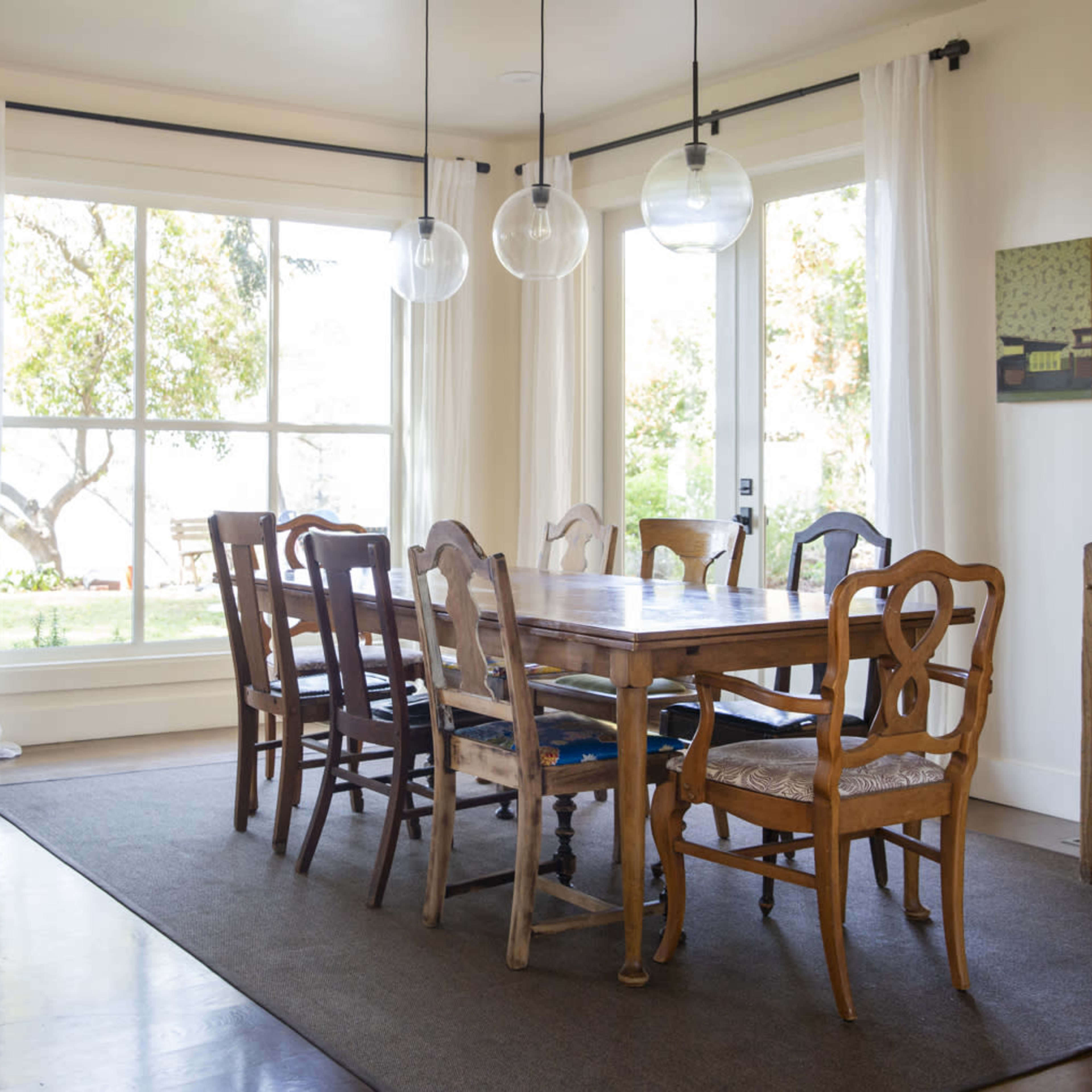A dining room features a large wooden table surrounded by eight chairs, with large windows letting in natural light and a plant in the corner.