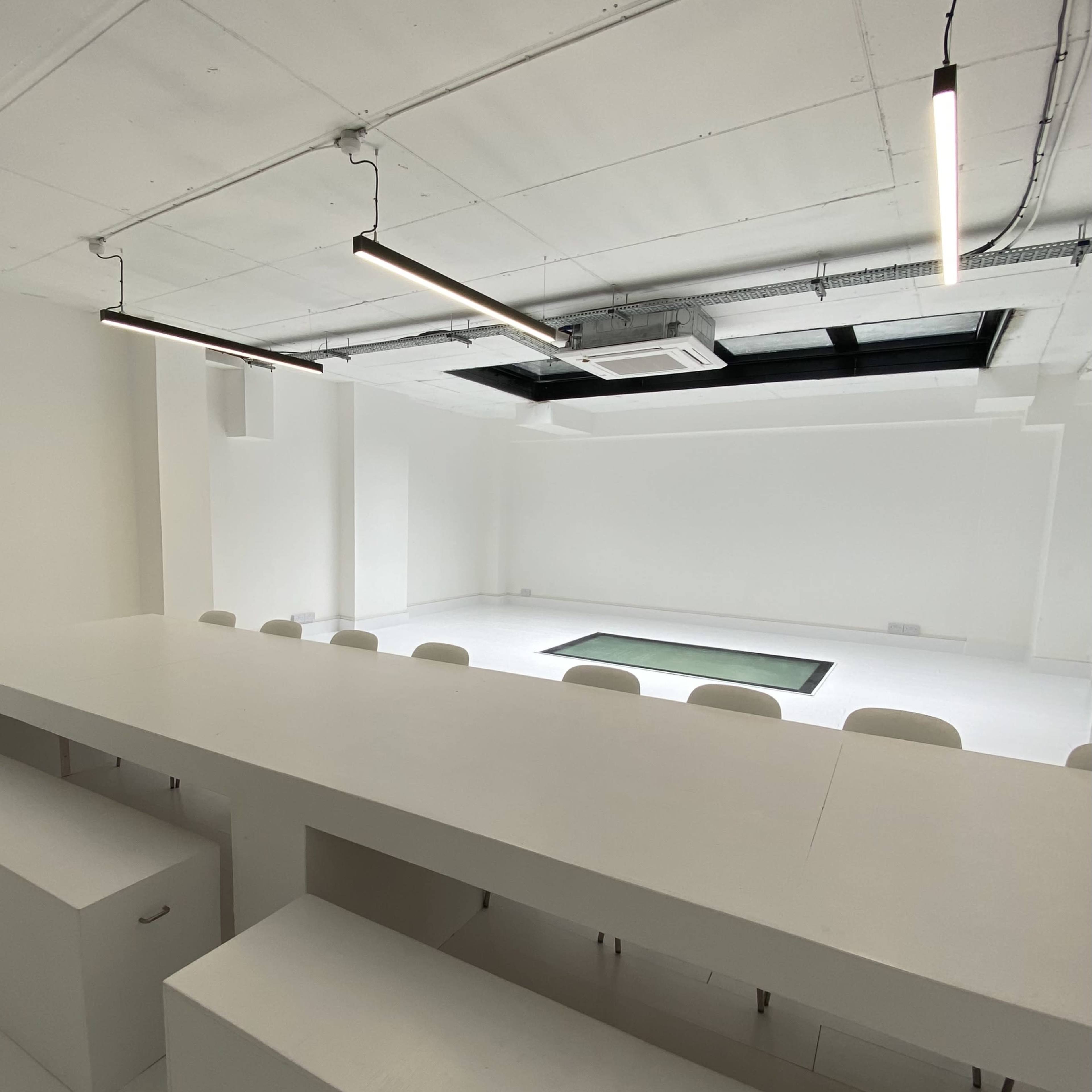 A minimalist, bright room with a long white table and chairs facing an empty space with a green mat on the floor.