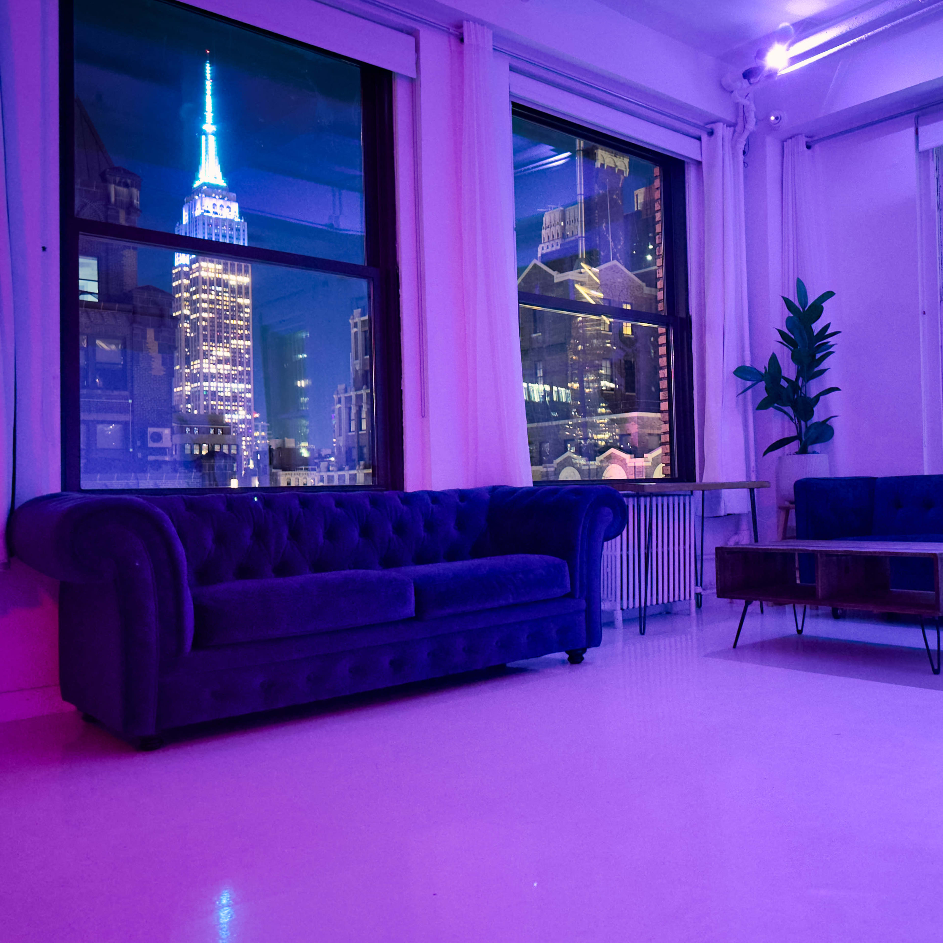A modern interior with two sofas facing a coffee table, and large windows revealing a city skyline featuring the Empire State Building illuminated at night.