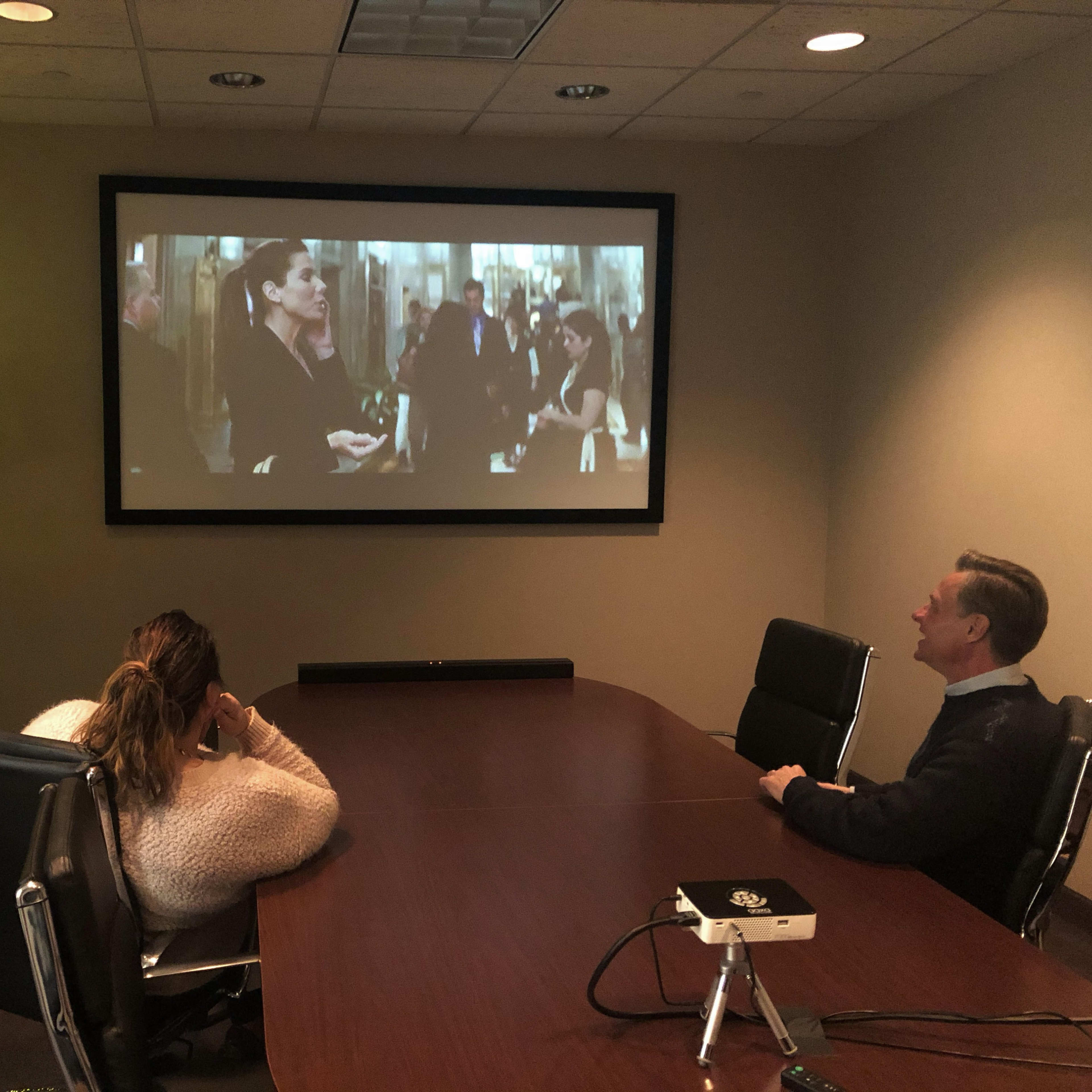 Two people sit at a conference table watching a movie on a large screen in a dimly lit office.