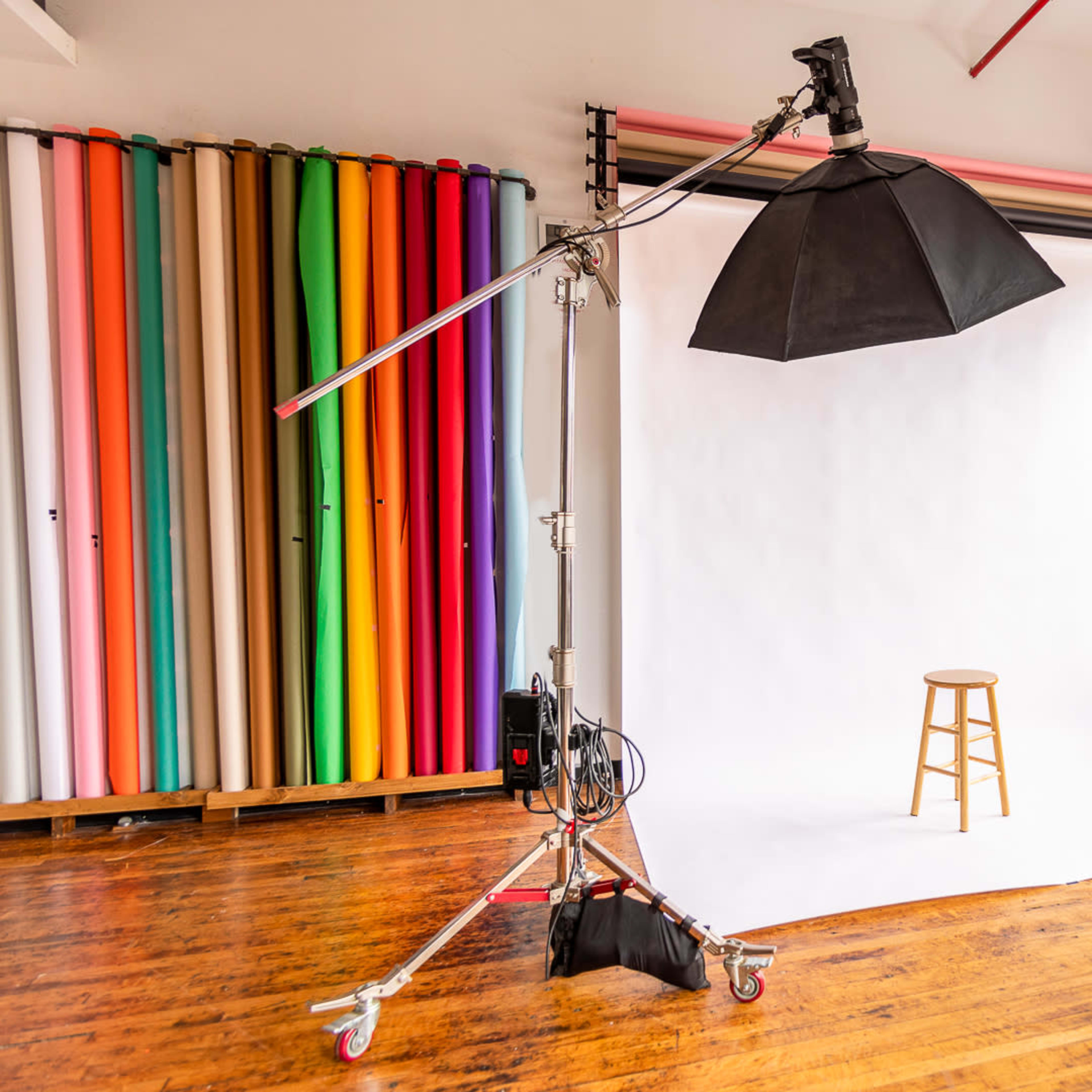 A photography studio with a white backdrop, a wooden stool, and various colored rolls of paper in the background.