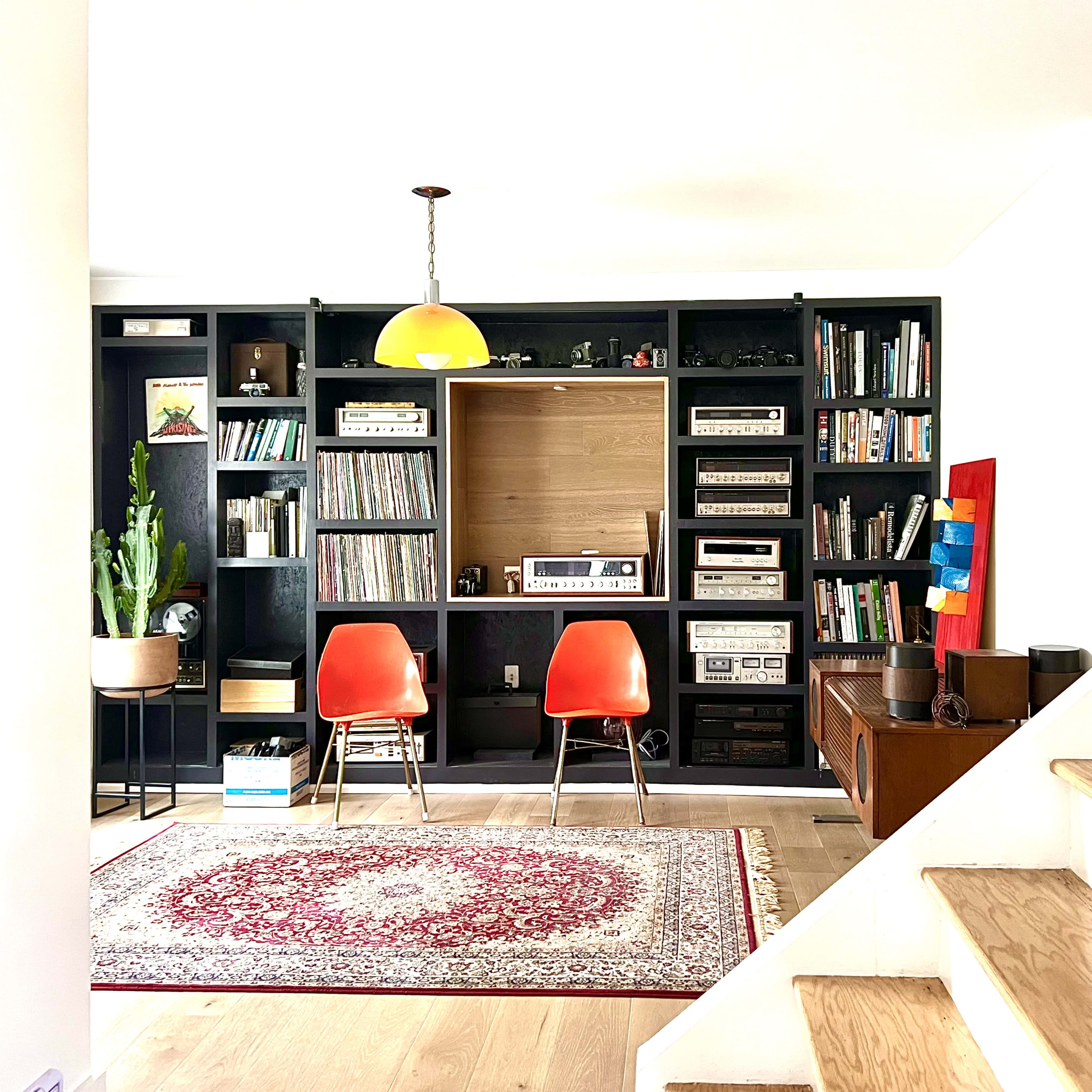 A living room with a bookshelf filled with records and equipment, two orange chairs, a rug on the floor, and a staircase on the right.